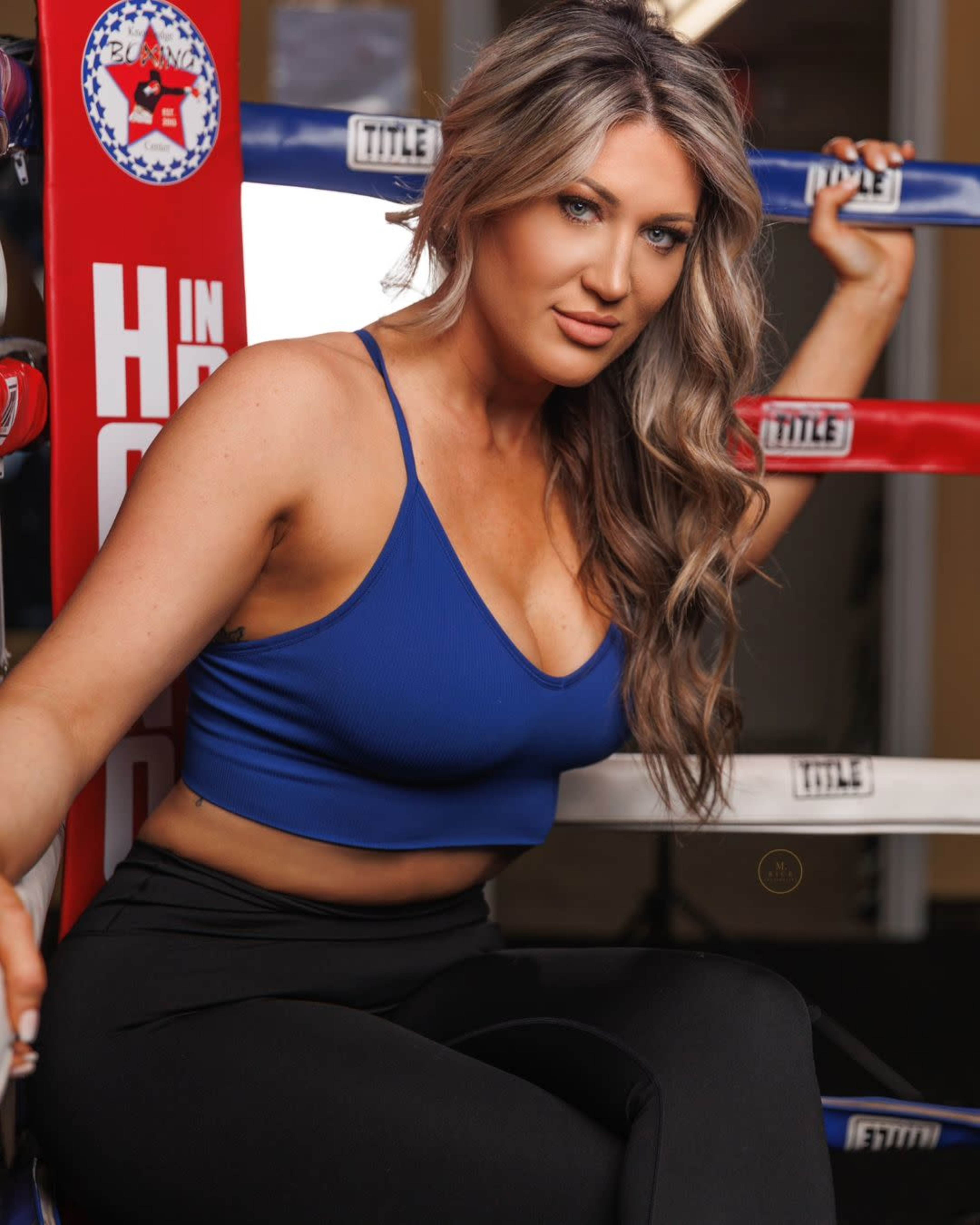 A woman in a blue tank top and black leggings poses on a boxing ring with red and blue corner posts.