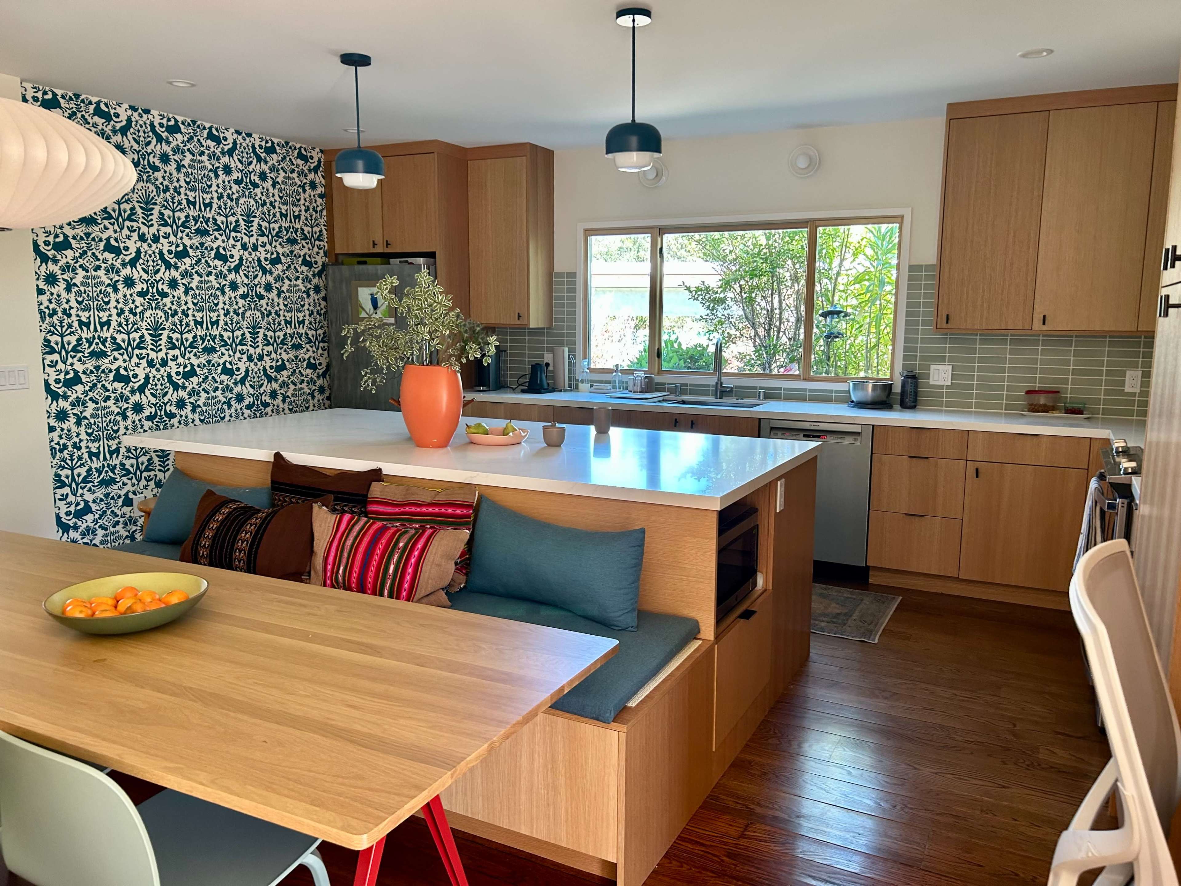 A modern kitchen features wooden cabinetry, a white island with seating, and a dining area with colorful cushions, alongside a large window overlooking greenery.