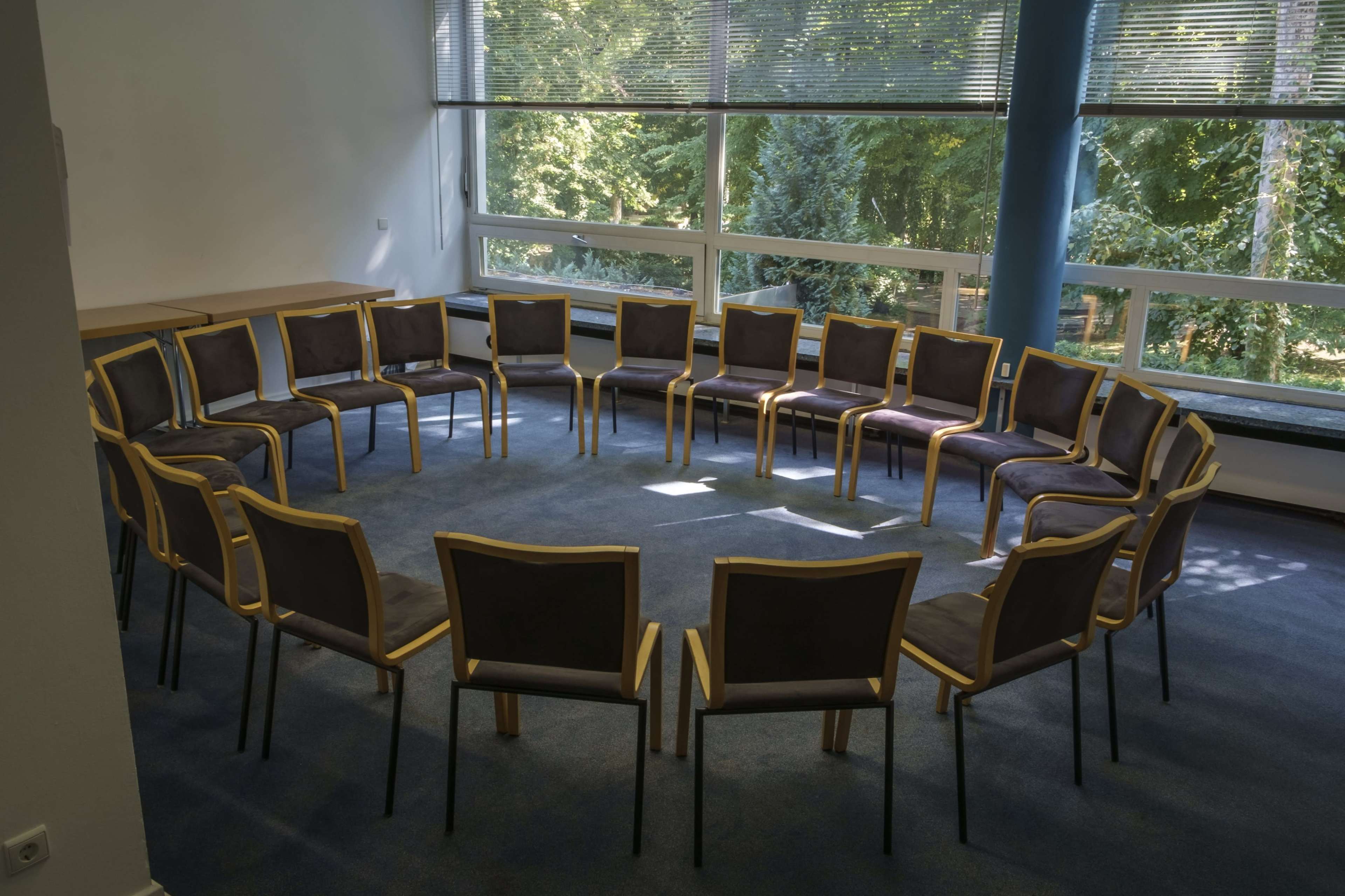 The image shows a circular arrangement of empty chairs in a room with large windows overlooking greenery.
