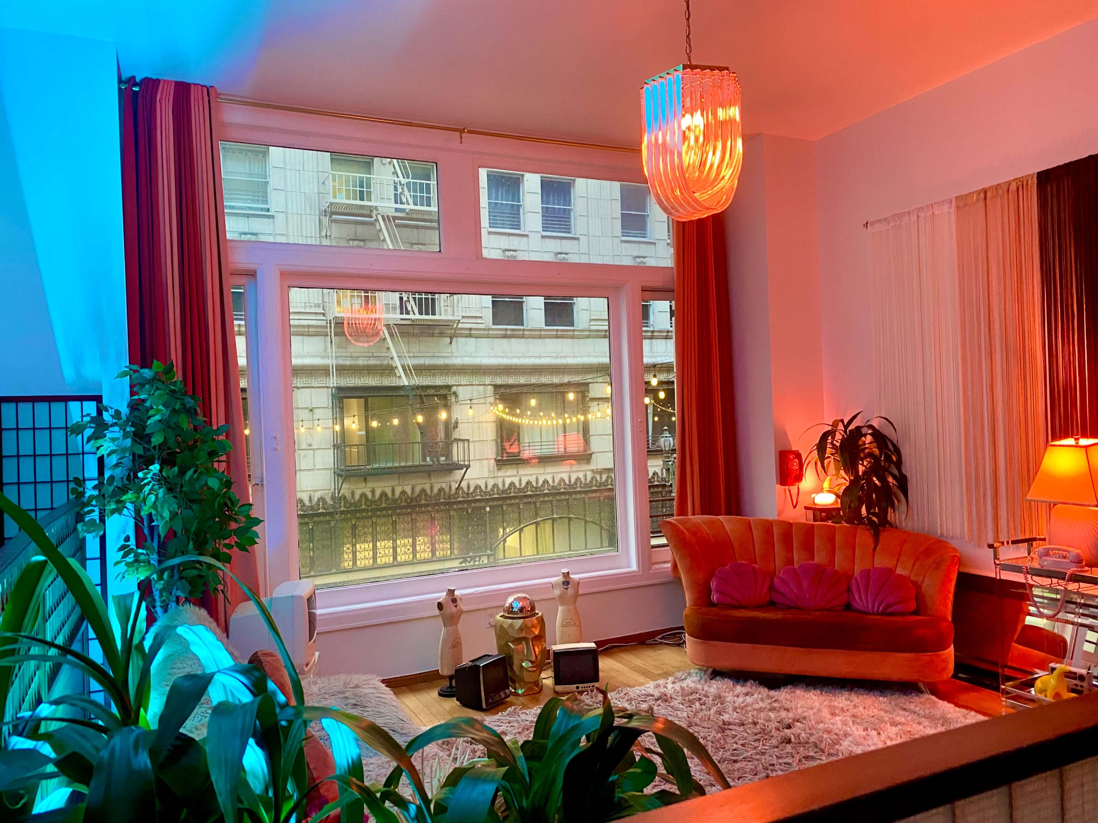 A cozy living room with a large window showcasing a view of a city street, featuring a vintage orange sofa, decorative plants, and warm lighting.