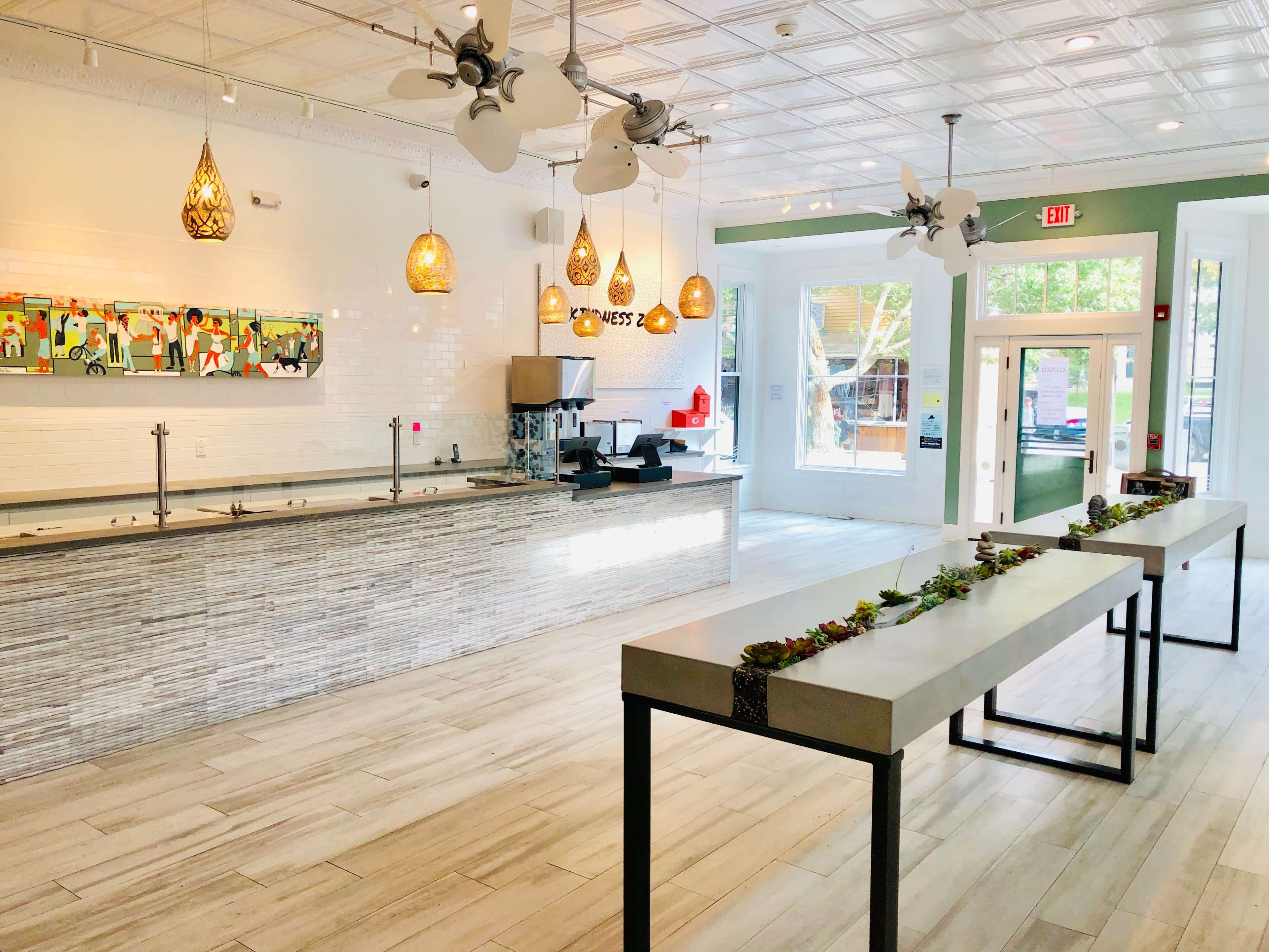 A modern, brightly lit interior of a café with a long counter and decorative pendant lights, featuring two long tables with planters along the center.