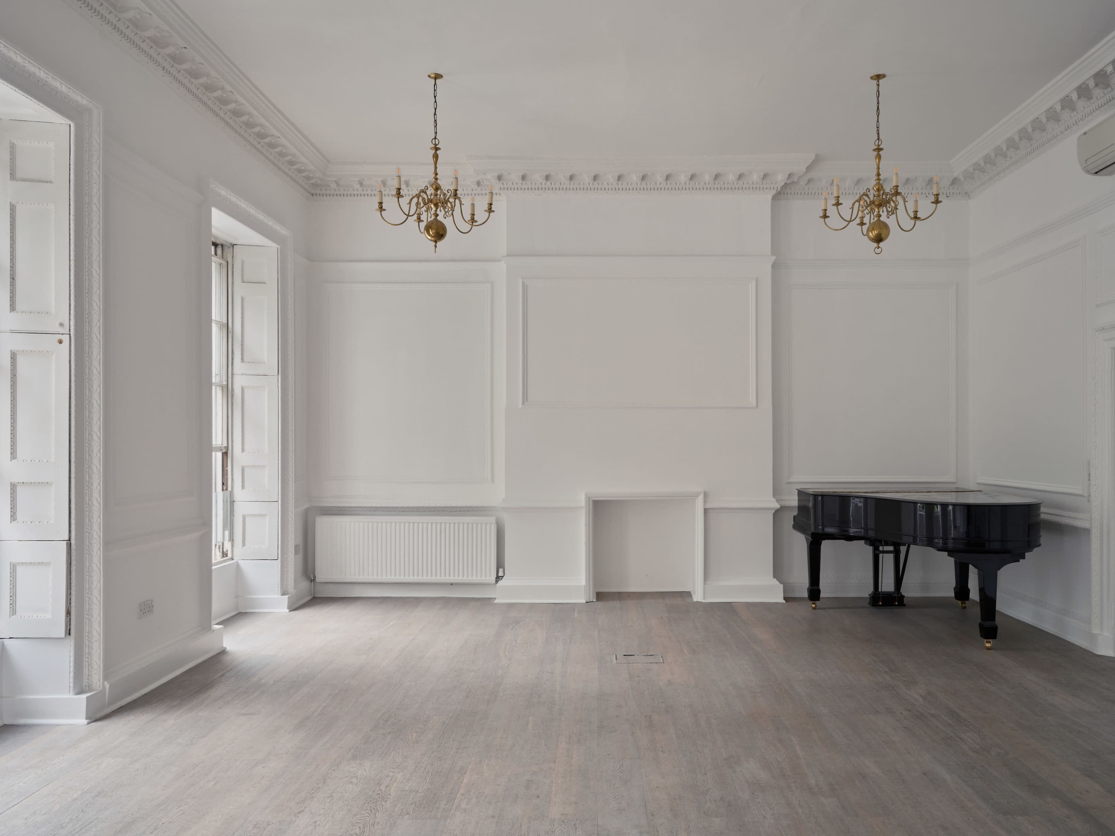 The image shows a spacious, empty room with white walls, wooden flooring, two chandeliers, a black piano in the corner, and large windows.