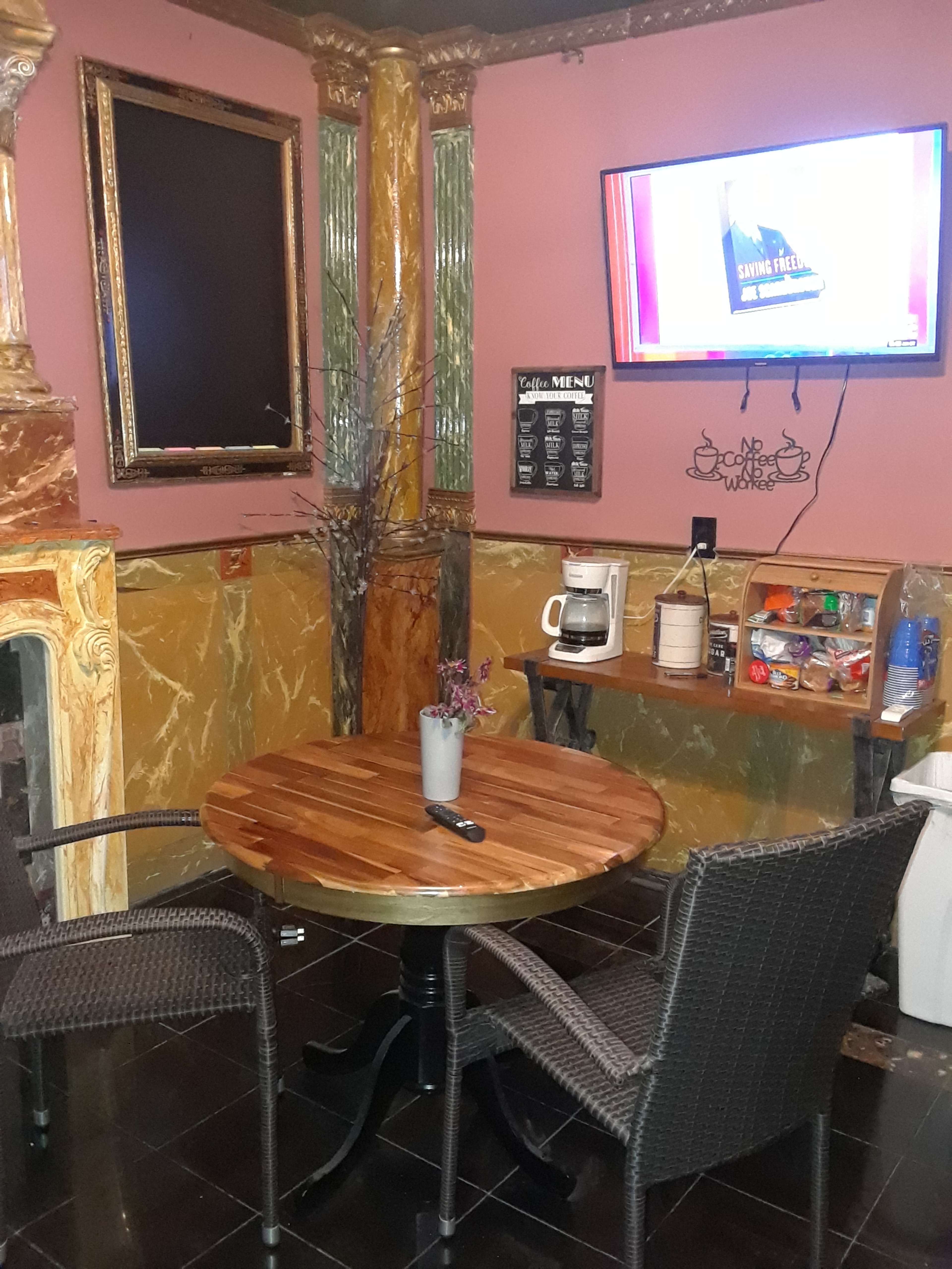 The image shows a small seating area with a wooden table, two wicker chairs, a coffee maker, snacks, and a TV mounted on the wall.