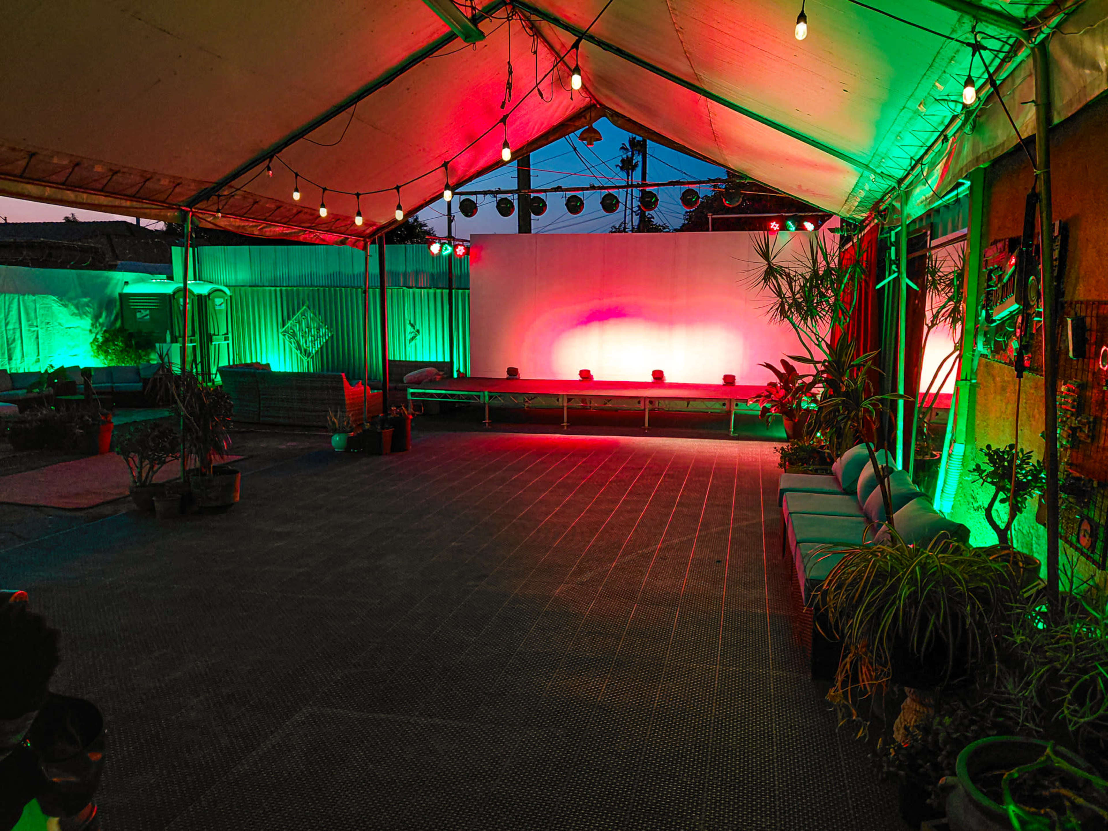The image shows a decorated outdoor tent with string lights and colored stage lights illuminating a seating area and performance space.