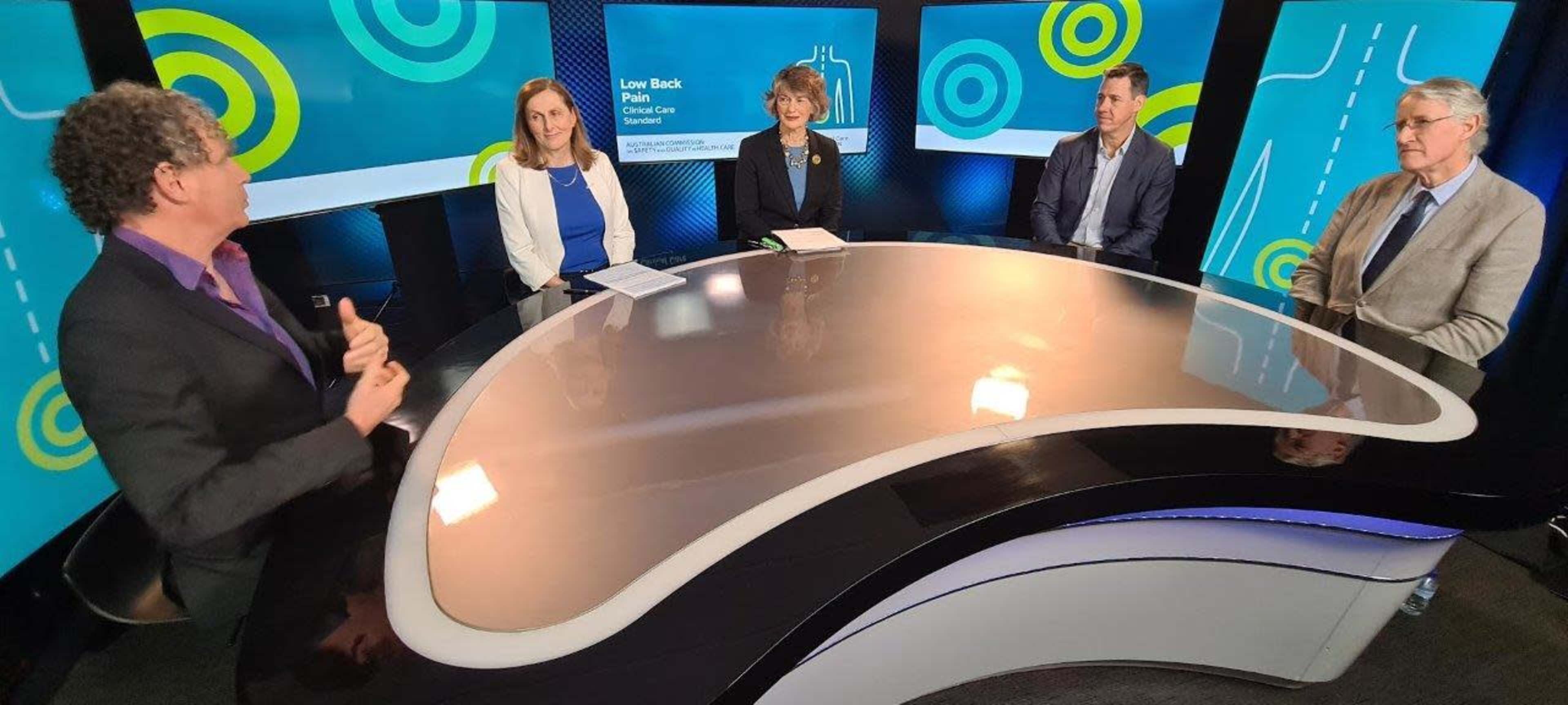 A group of five people is seated around a modern, curved table in a studio setting, discussing a topic related to a "Lawn Back Plan" presentation, with graphics in the background.