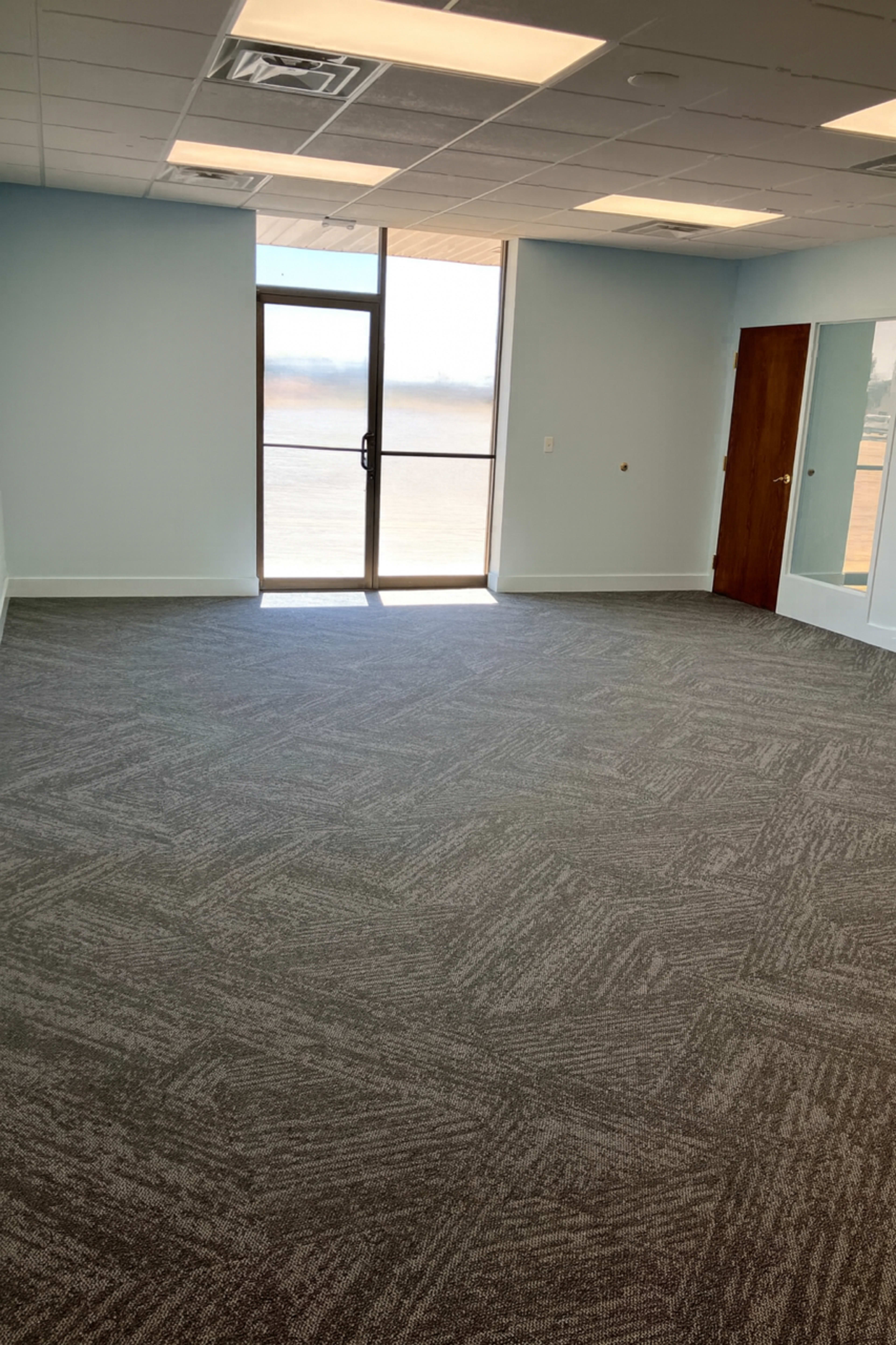 The image shows an empty office space with gray carpet, a large window, and a wooden door leading to a balcony or outdoor area.