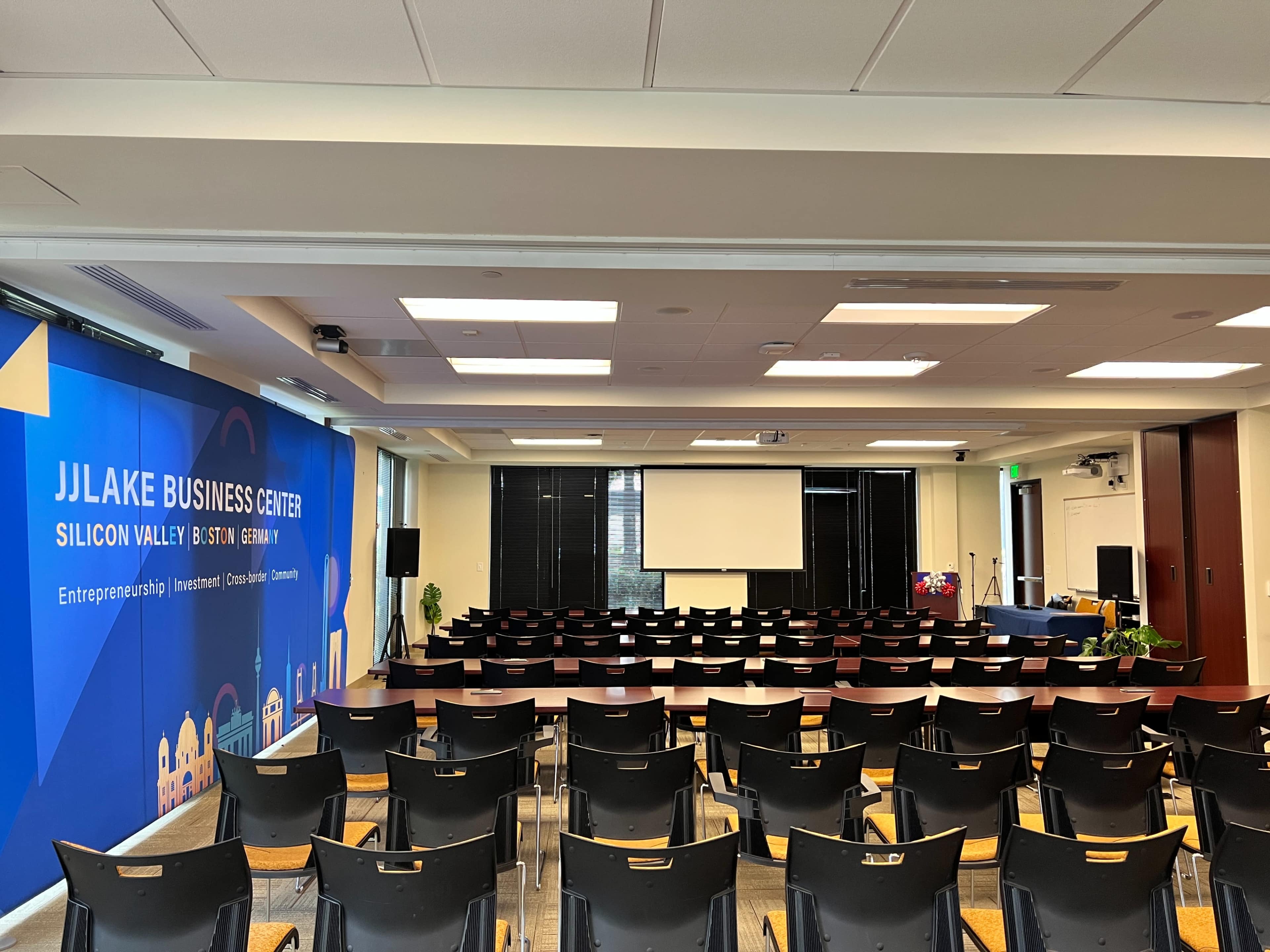 A conference room set up for a presentation, featuring rows of black chairs facing a projector screen and a large wall display for the JJLake Business Center.
