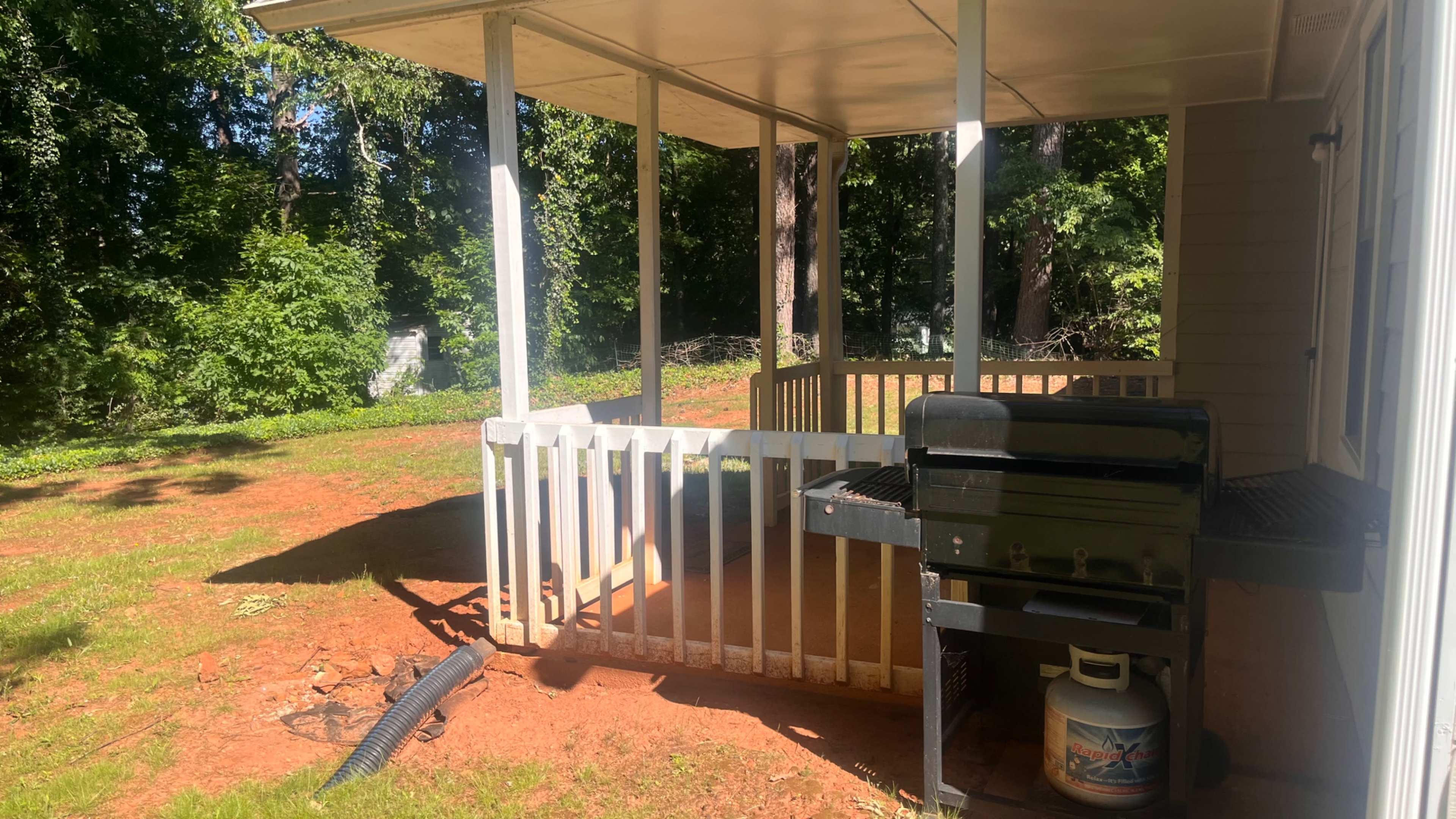 The image shows a covered porch with a black gas grill beside a white wooden railing, set in a yard with sparse vegetation and a few trees in the background.