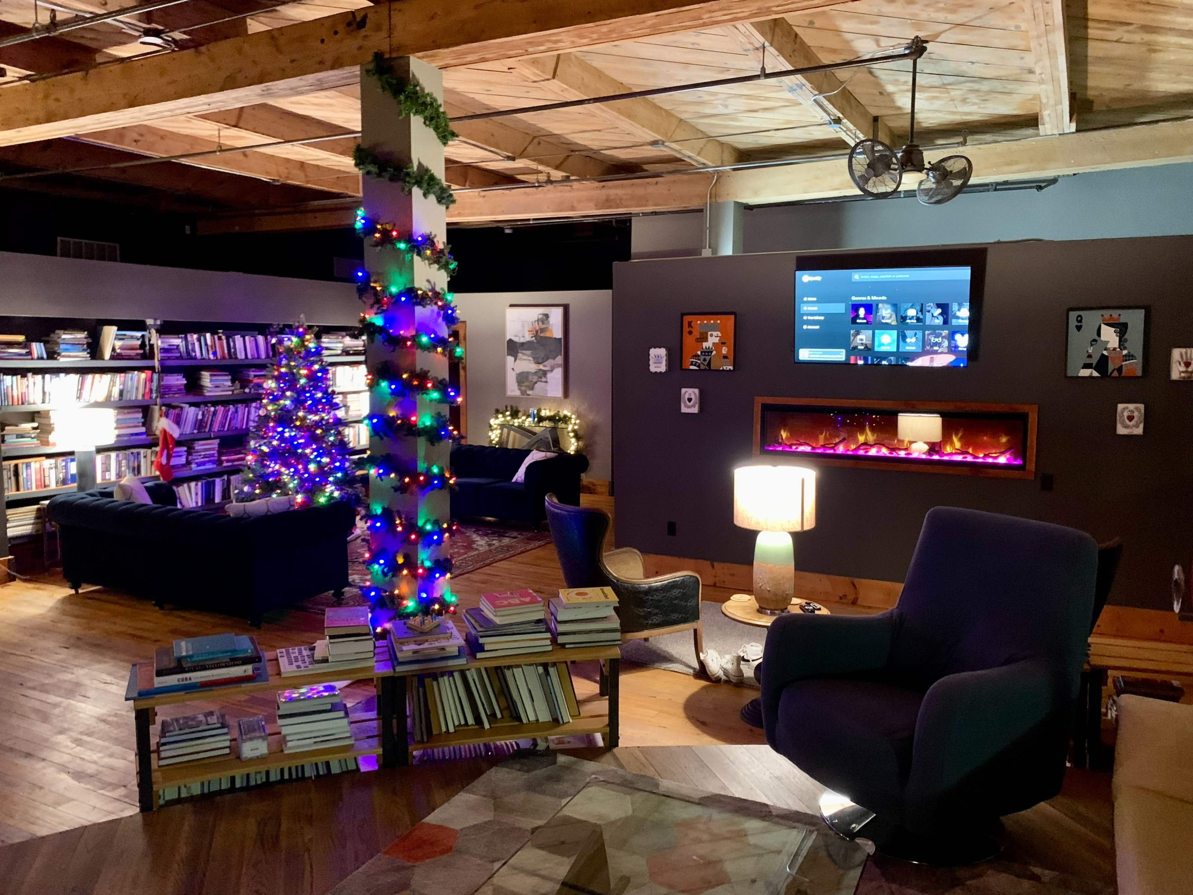 A cozy library area with bookshelves, a Christmas tree decorated with lights, and a lounge space featuring a large chair and a modern television setup.