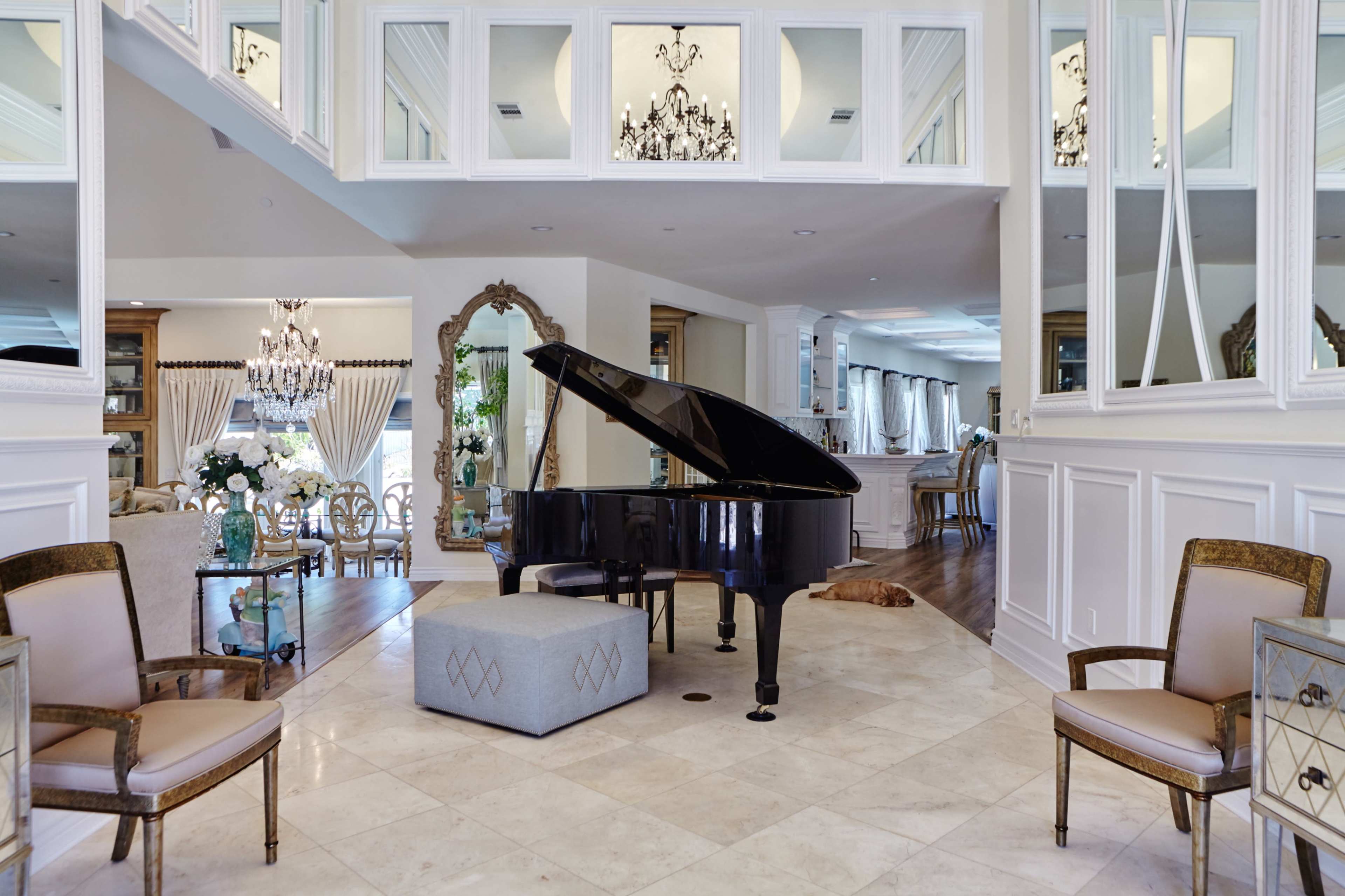The image shows an elegantly designed interior featuring a grand piano, a mirrored wall, and a chandelier, with light-colored tile flooring and ornate furnishings.