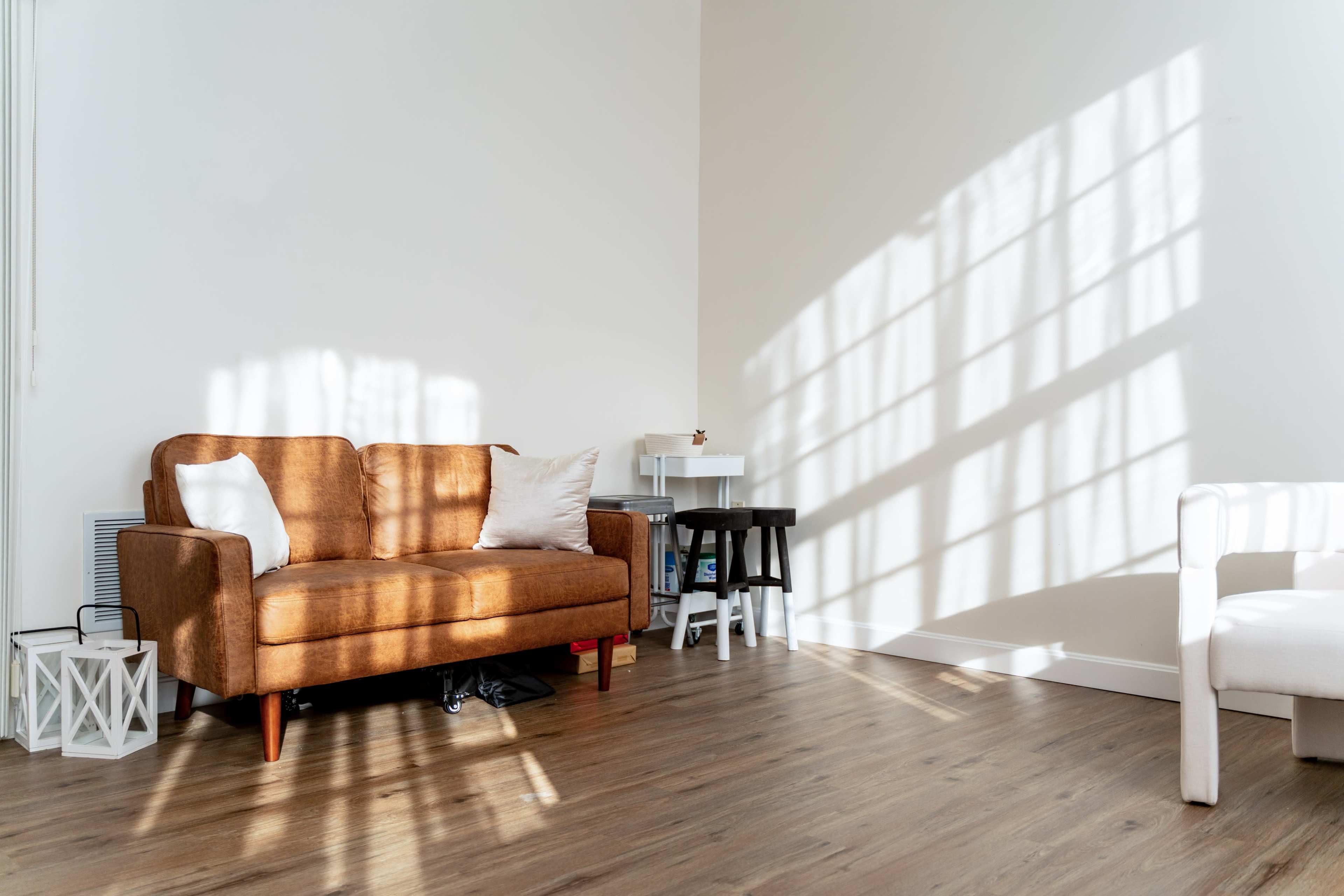 A brown sofa with a white pillow sits against a wall, casting shadows from nearby window panes on the floor.