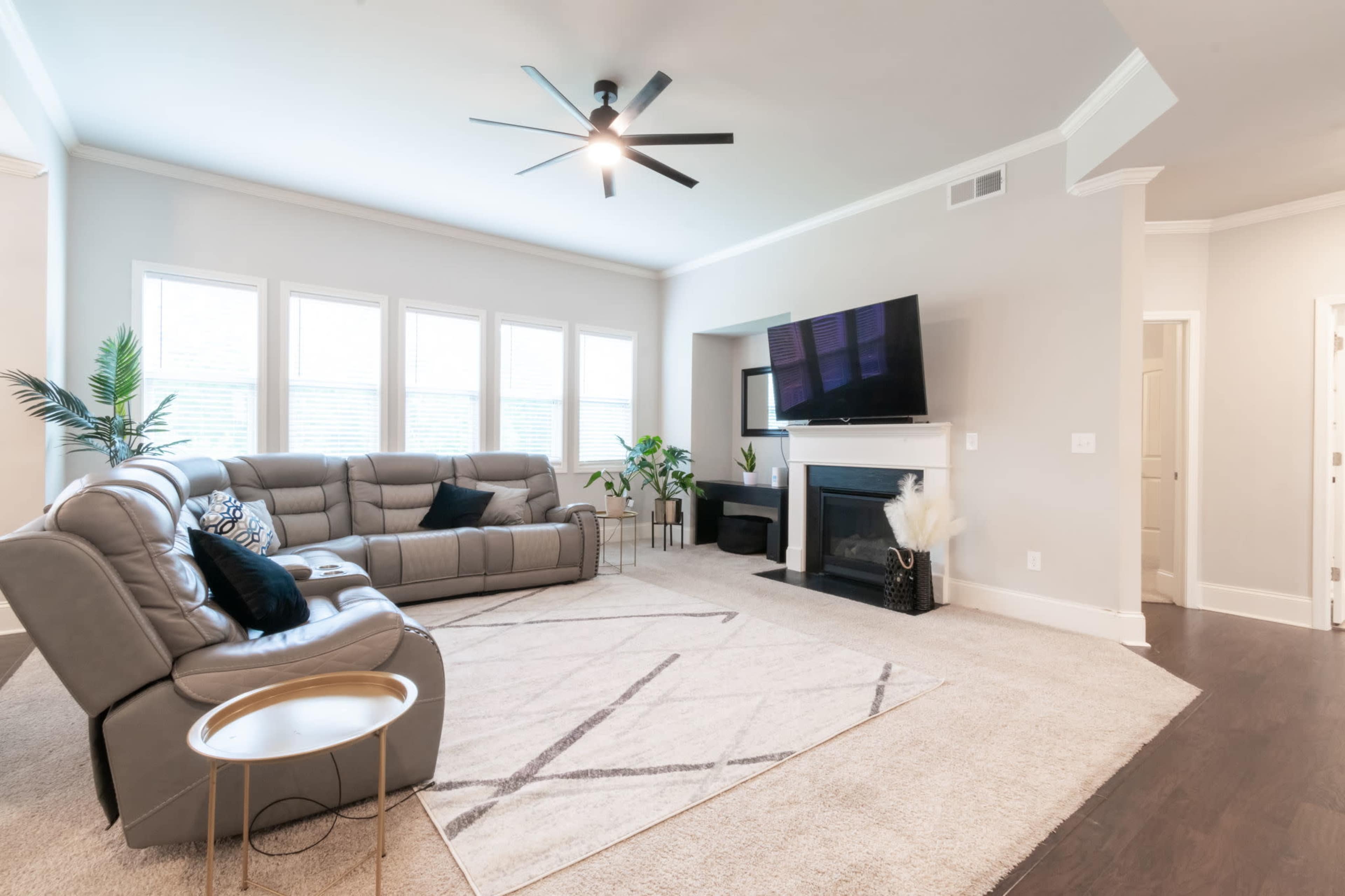 A modern living room featuring a sectional sofa, a coffee table, a large flat-screen TV mounted above a fireplace, and several plants near the windows.