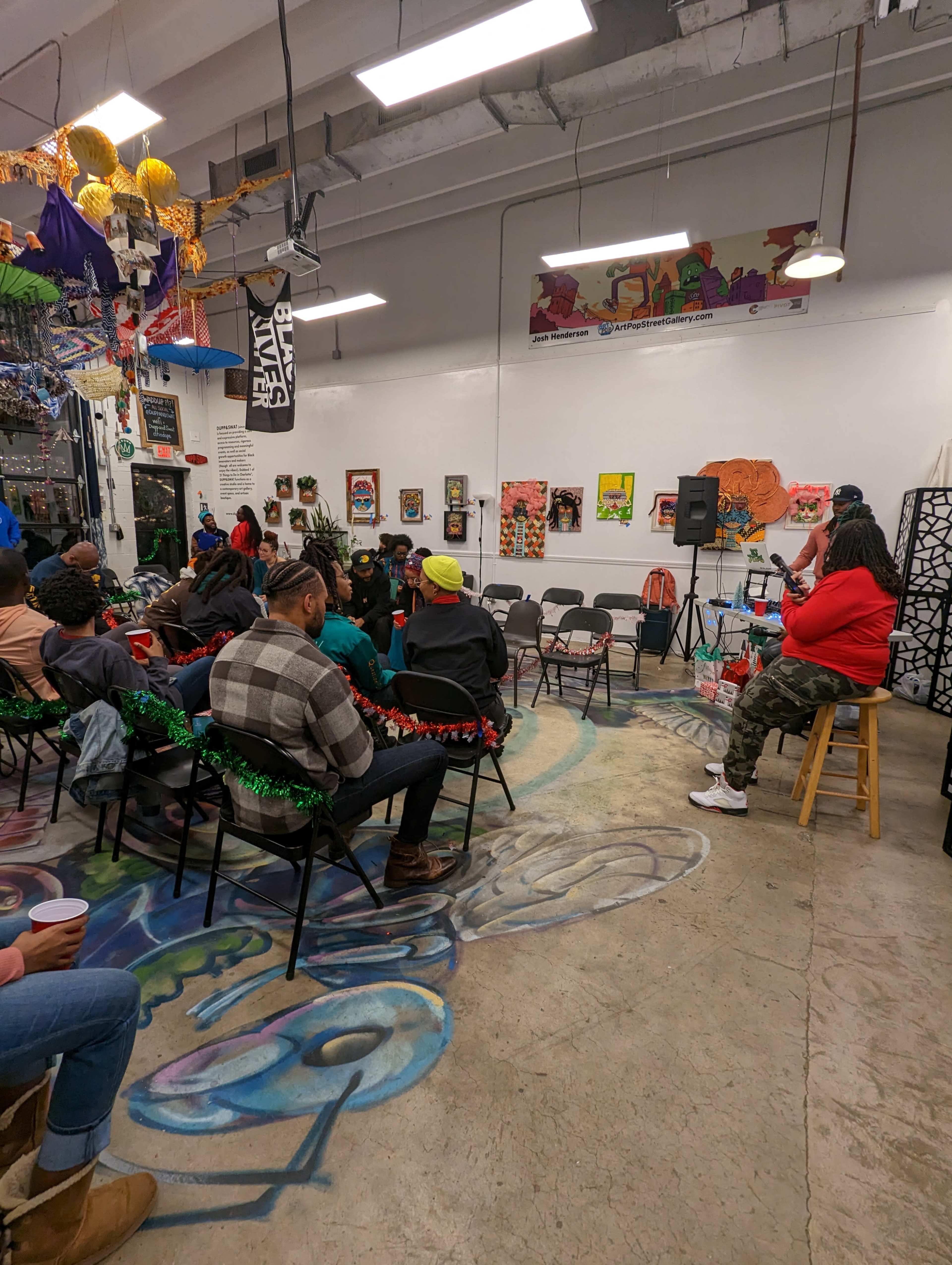 A group of people sits on chairs in a colorful room decorated with various artworks and a stage for a speaker.