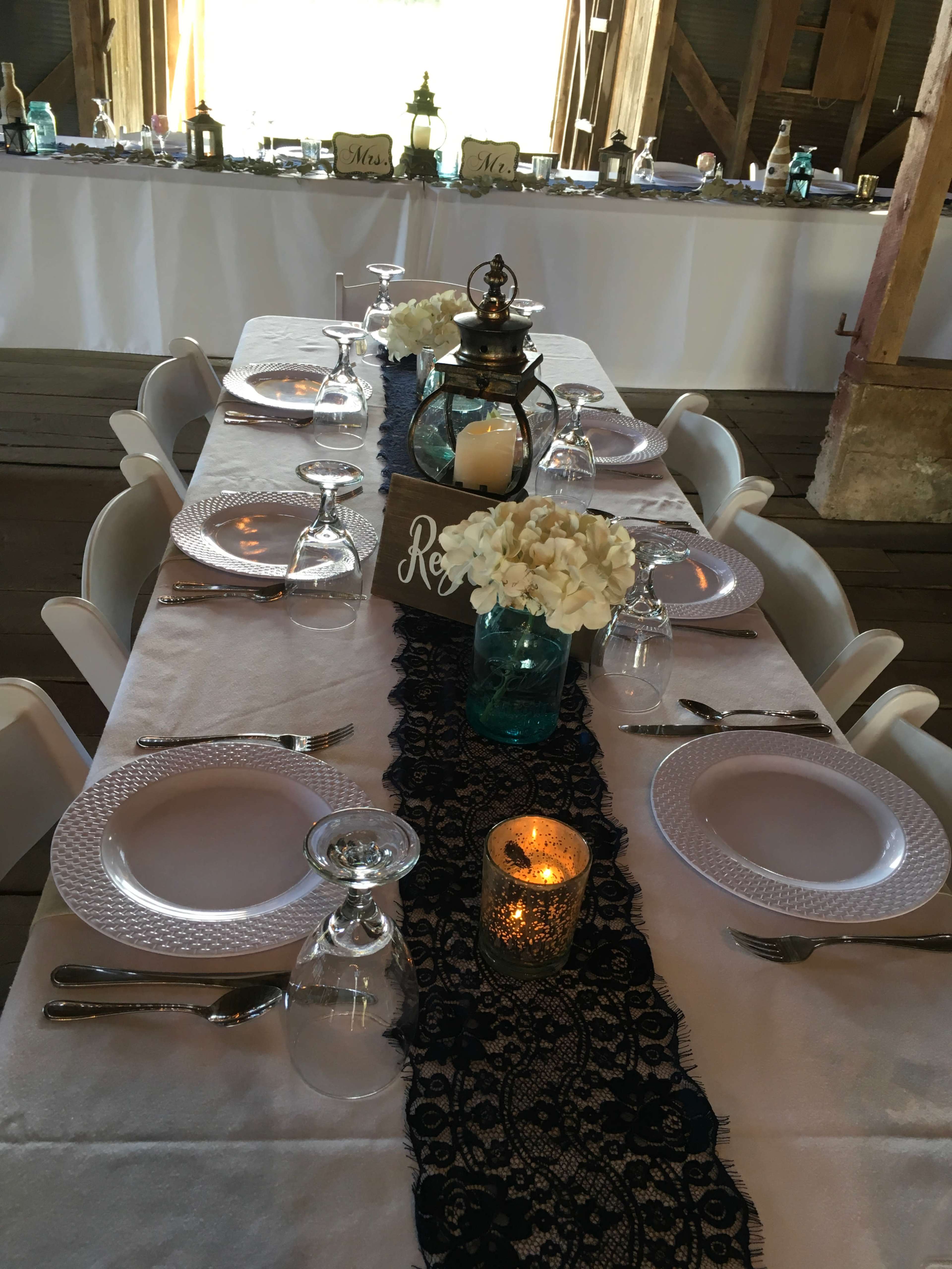 A long dining table is set with white plates, glasses, and centerpieces of flowers and candles, all arranged atop a dark lace runner in a rustic venue.