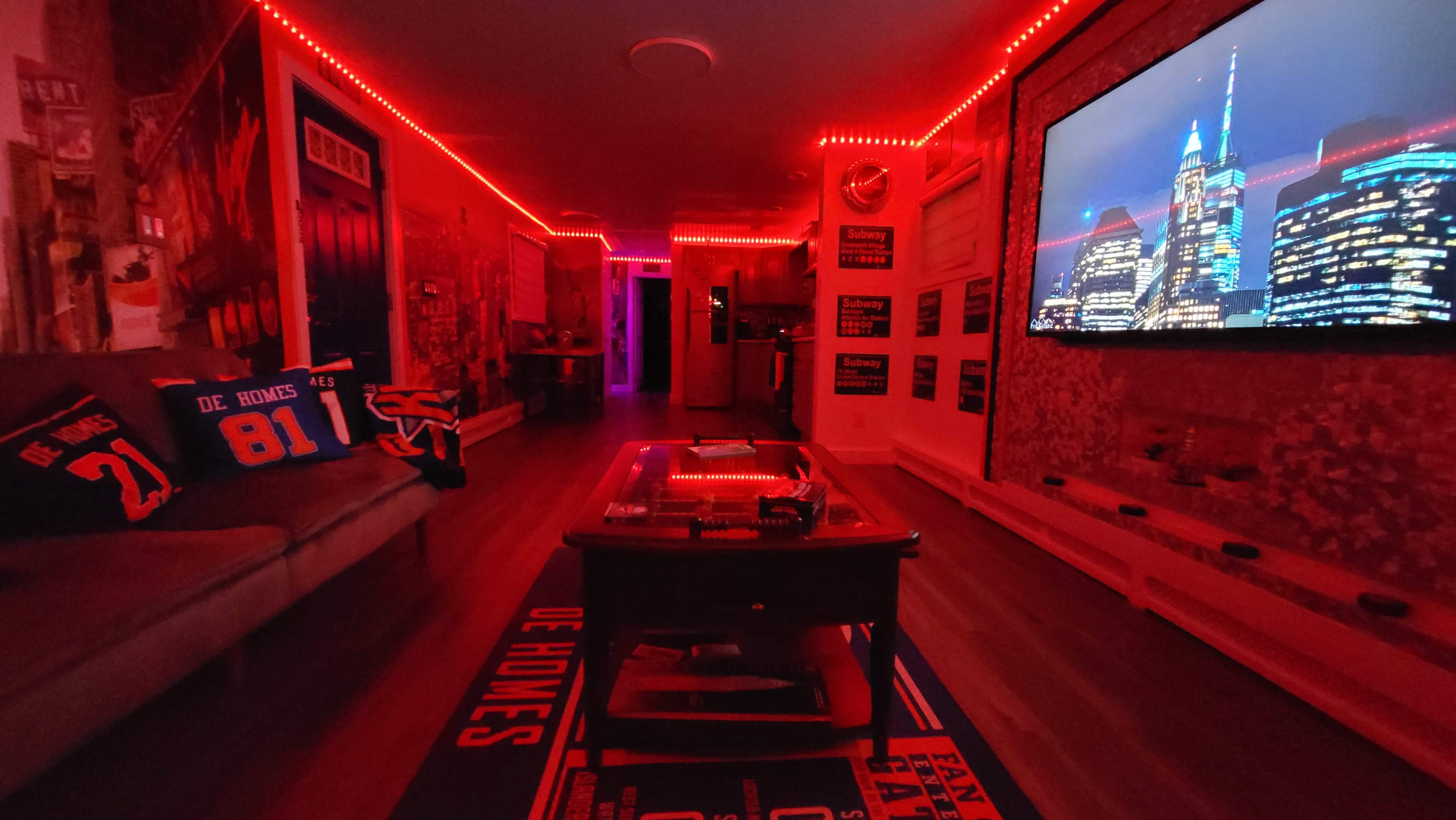 The room is illuminated with red LED lights, featuring a large screen displaying city skyline images, a couch with themed pillows, and a coffee table.