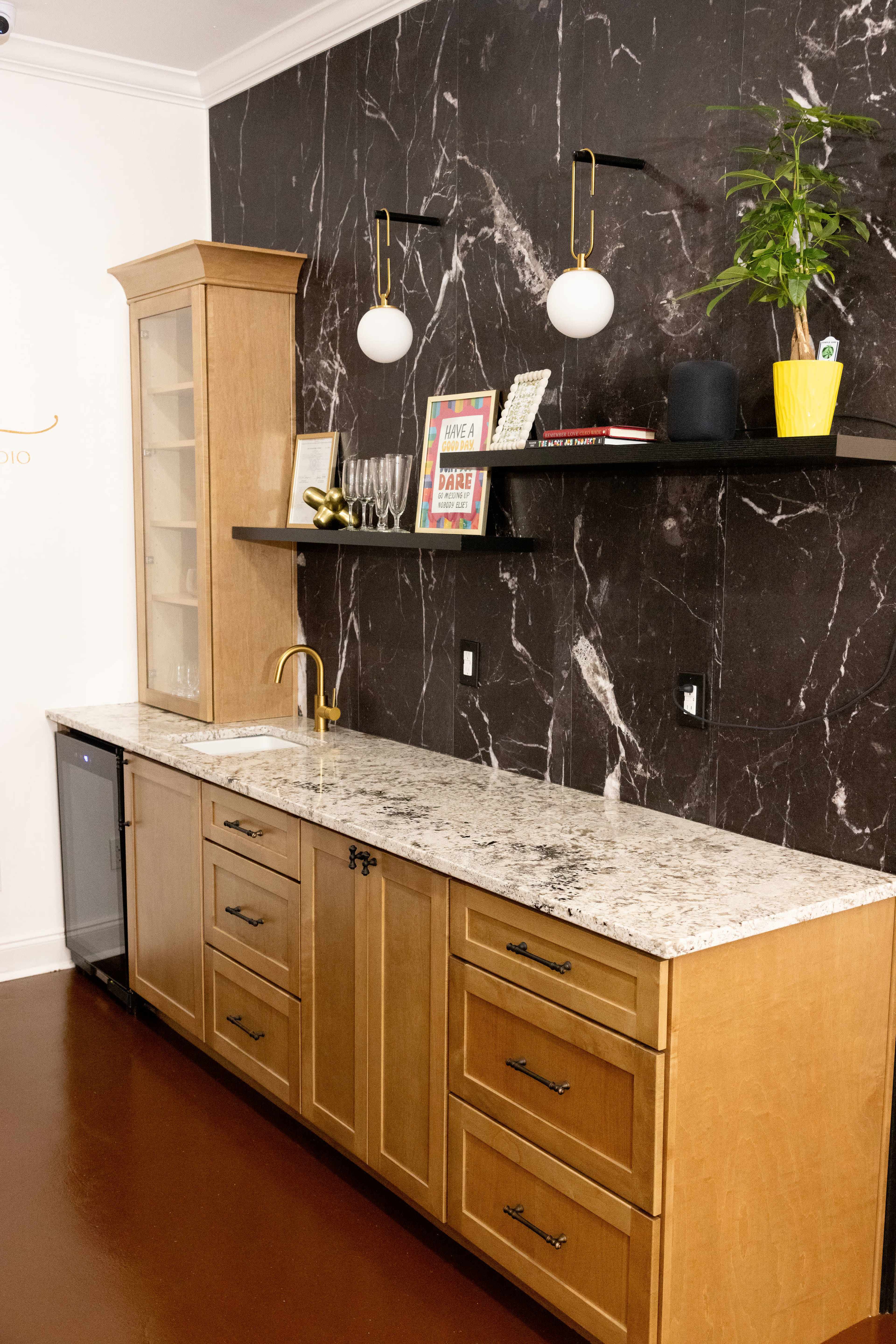 The image shows a modern kitchen area featuring a granite countertop, wooden cabinets, a glass-front cupboard, and a black marble accent wall.