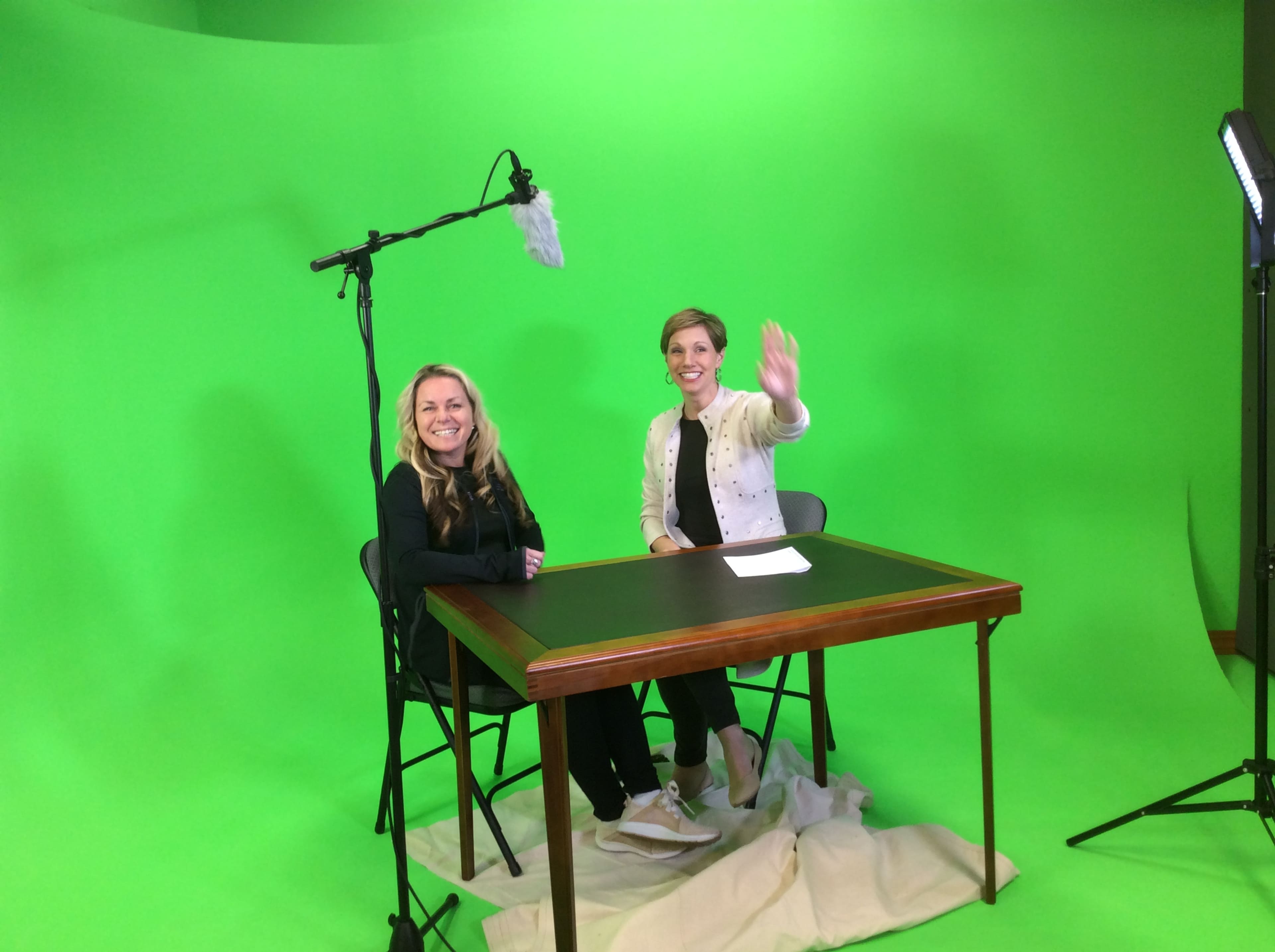 Two women are seated at a table in front of a green screen, with a microphone on a boom above them and a light set up to the side.