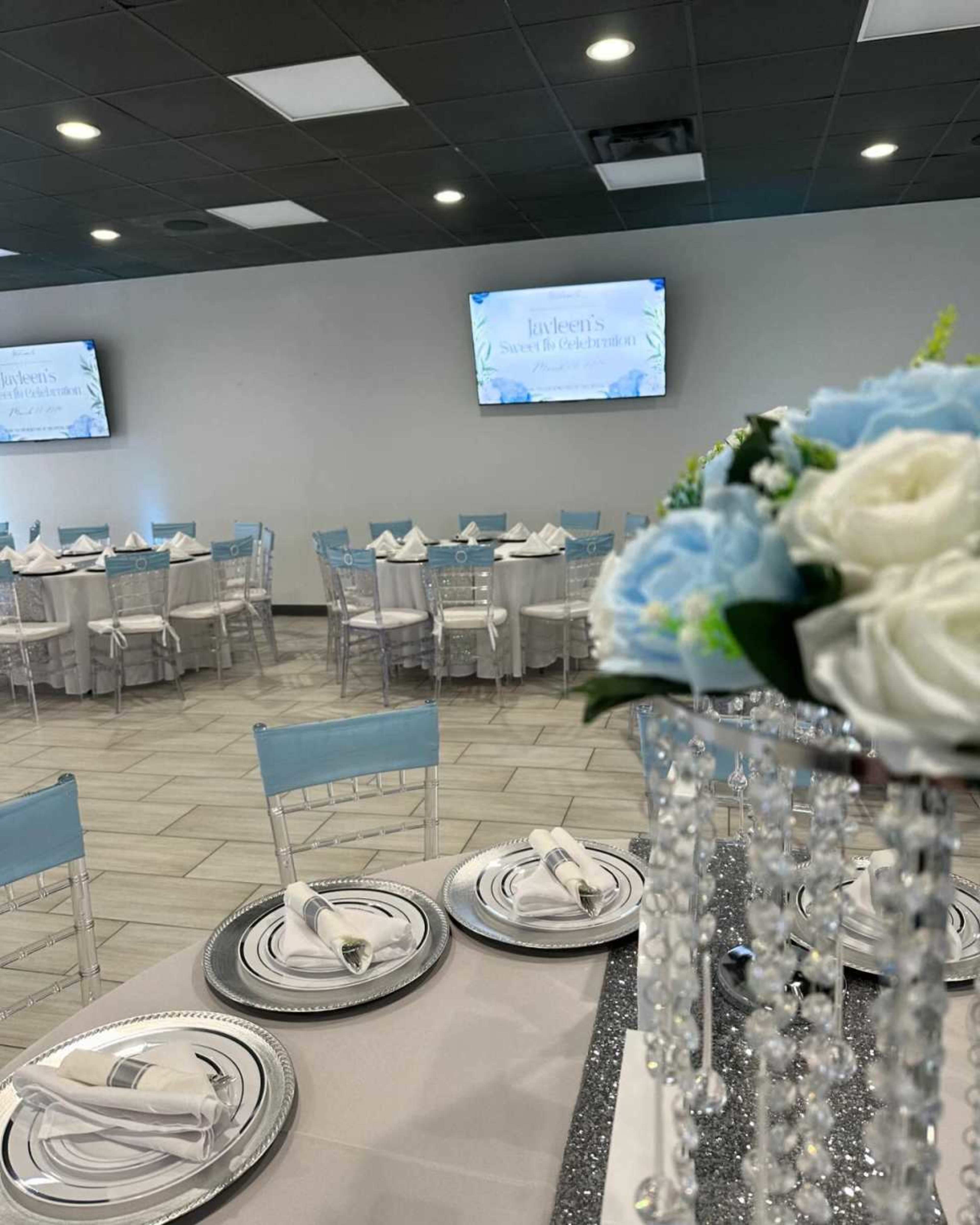 The image shows a decorated banquet hall with tables set for an event, featuring blue and white decor, elegant table settings, and screens displaying text in the background.