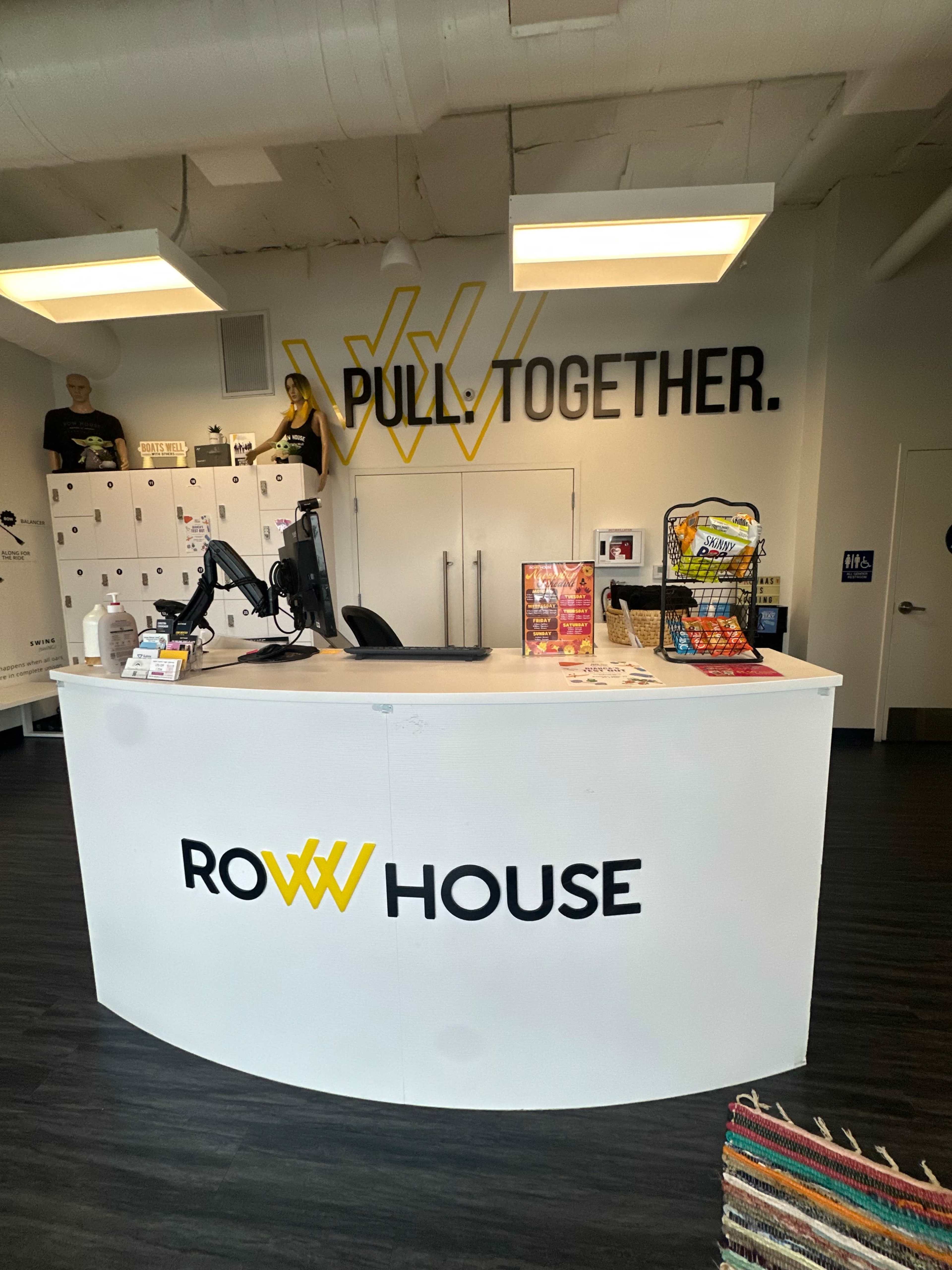 The image shows the reception area of a fitness studio called Row House, featuring a clean white desk with branding and promotional materials, along with lockers and a few decorative items in the background.