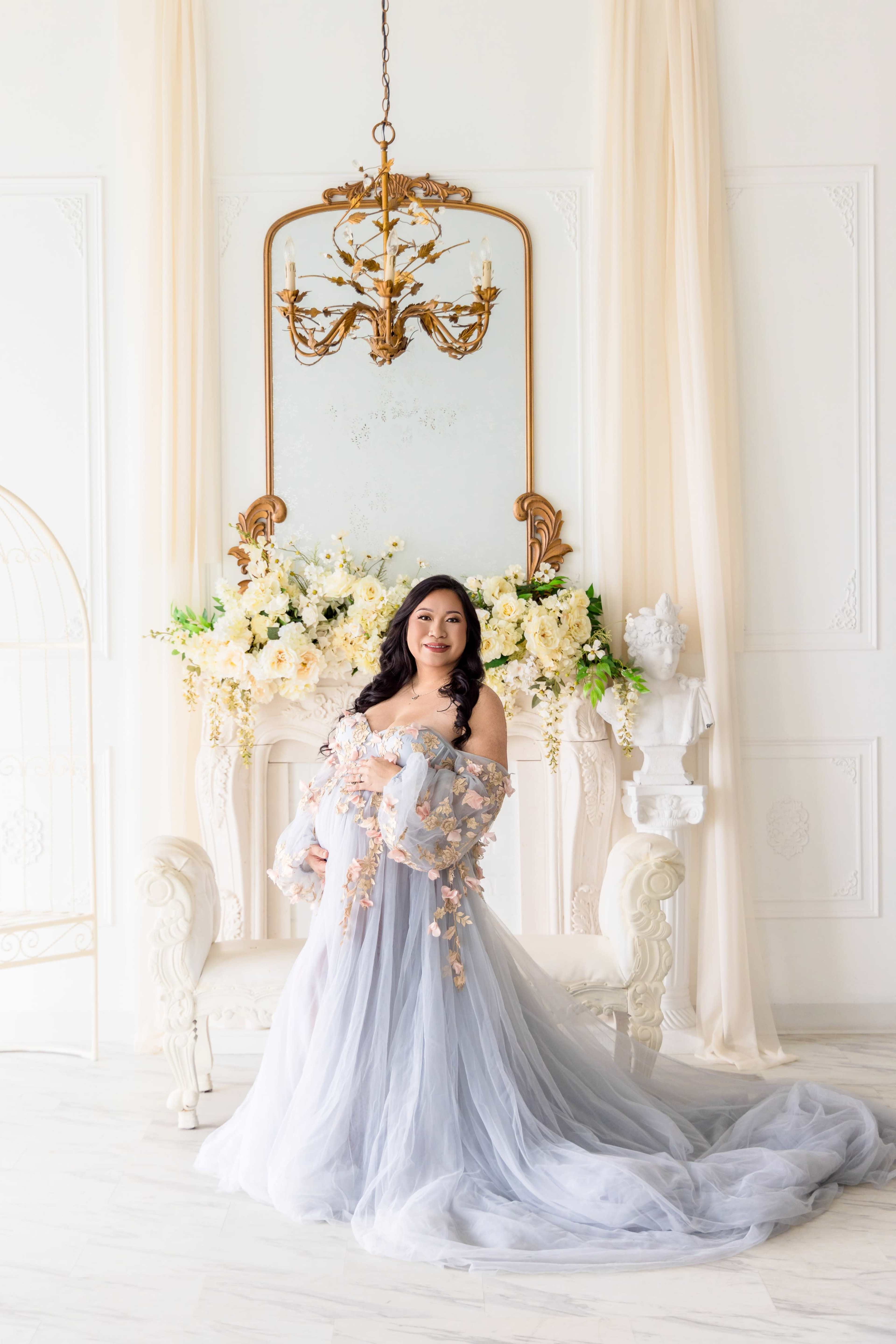 A woman in a flowing gray gown stands elegantly beside a floral-decorated fireplace in a bright, softly lit room.