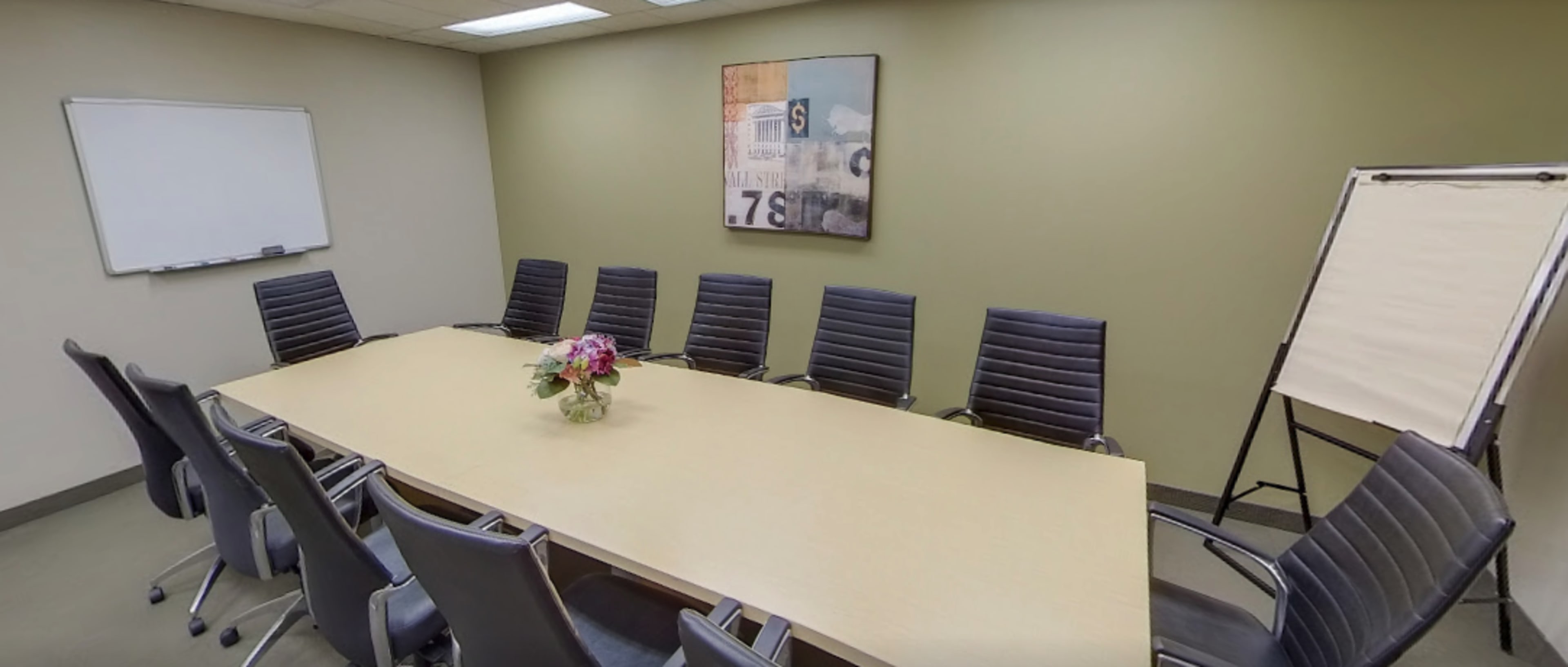 A conference room features a long table surrounded by eight black chairs, with a floral centerpiece and a whiteboard on one wall.