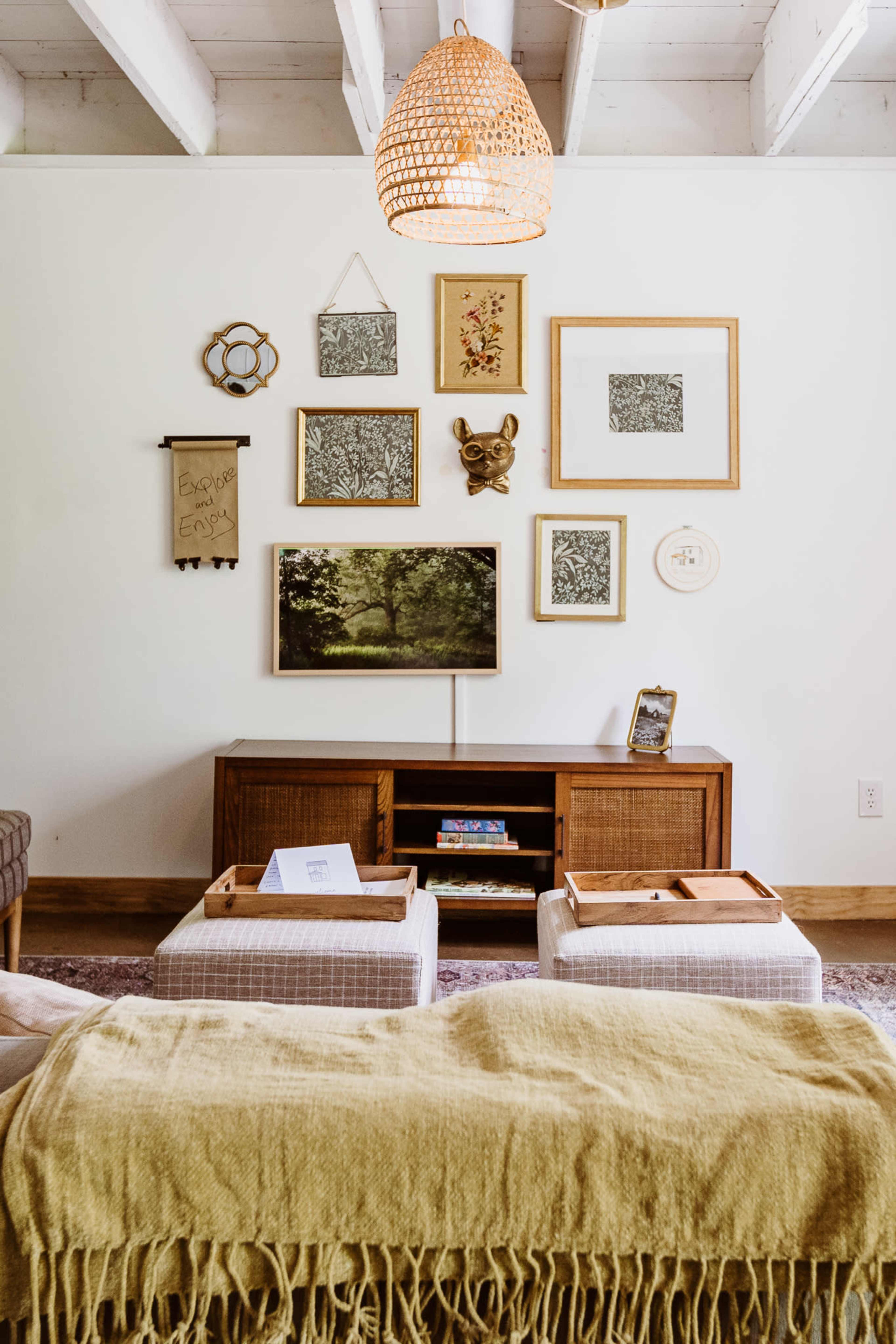 A living room with a wooden TV stand, decorative art on the wall, a woven light fixture, and a yellow throw blanket draped over a sofa.