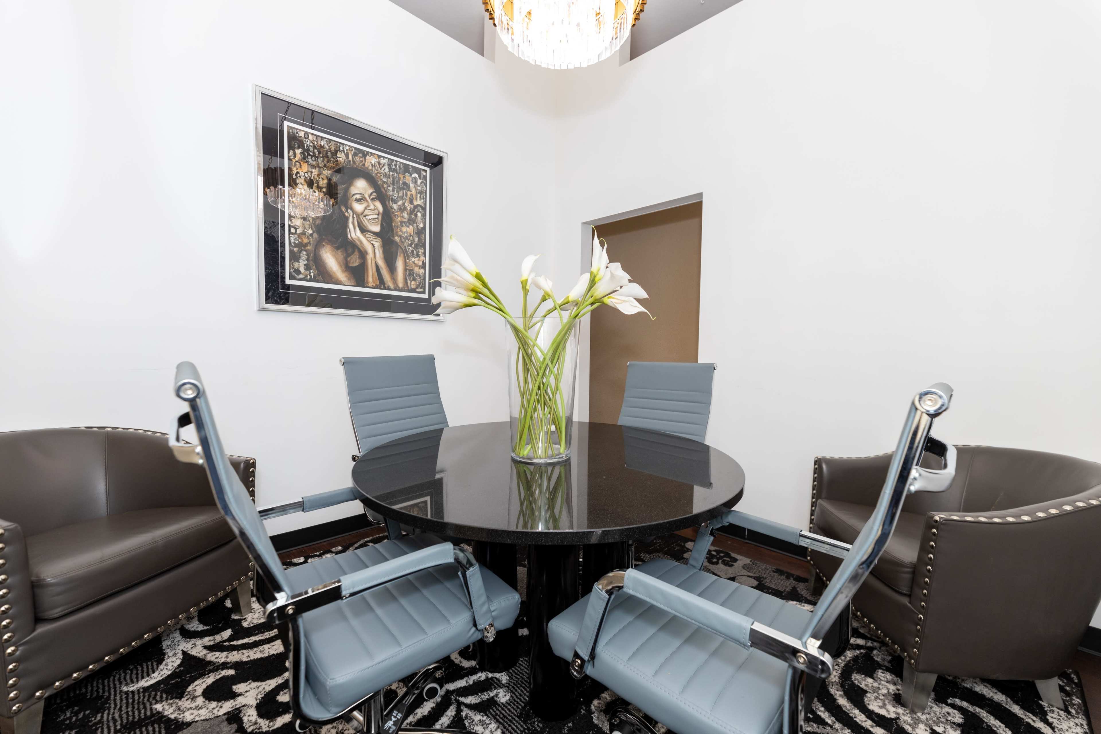 A modern conference room with a round black table surrounded by four blue and gray chairs and a large vase of white flowers on the table.