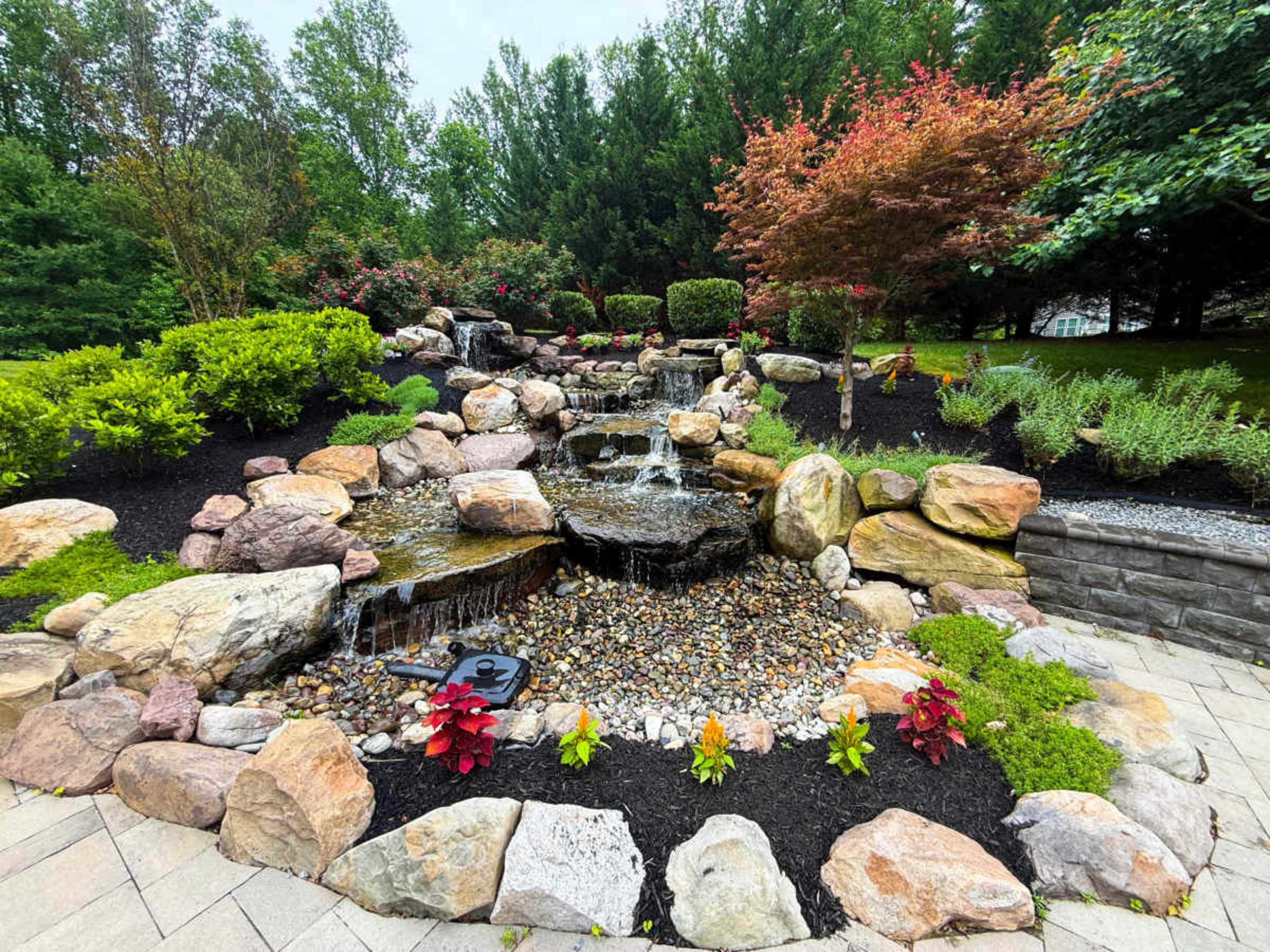 The image shows a landscaped garden featuring a cascading water feature surrounded by various rocks, shrubs, and colorful plants.
