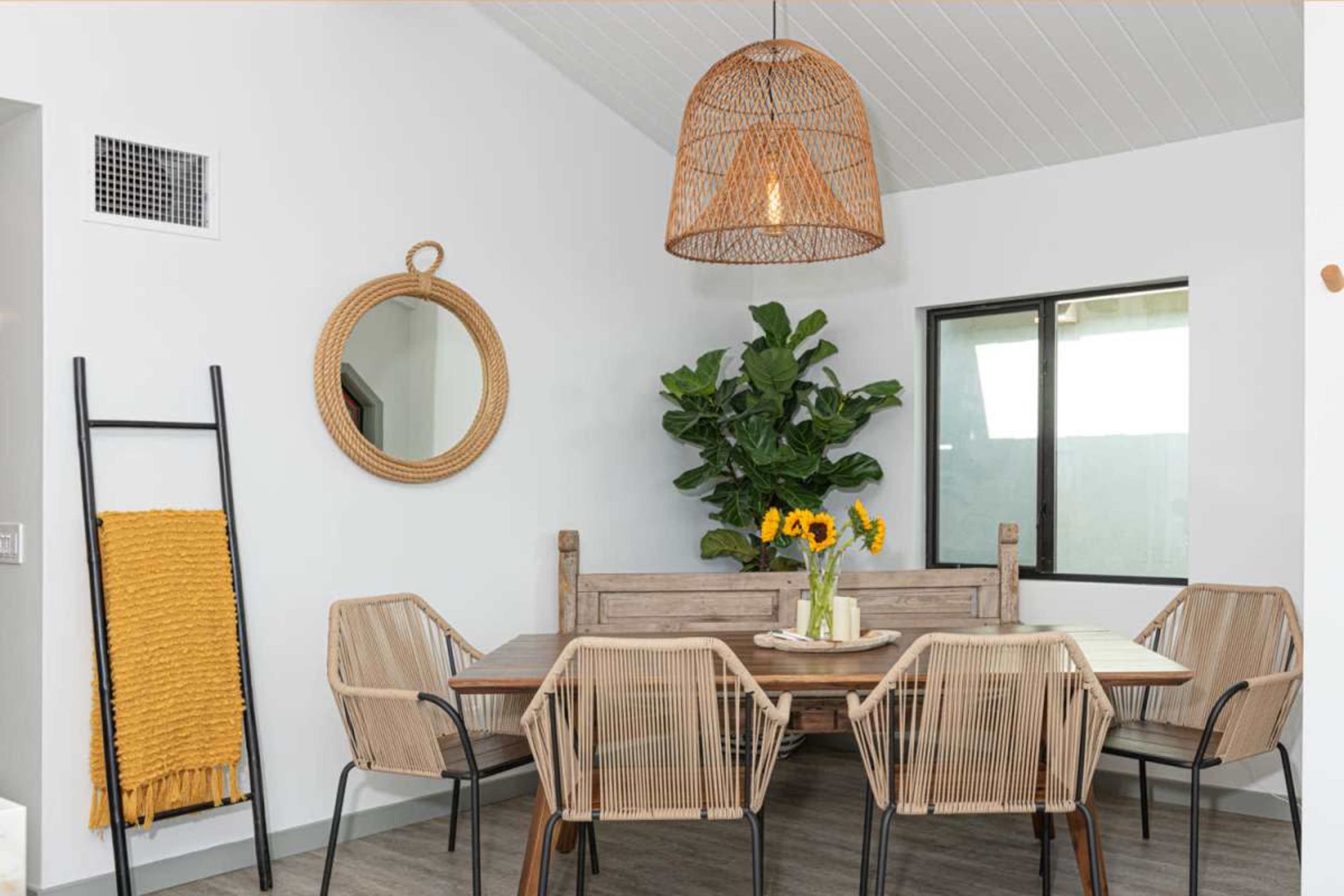 A dining area features a wooden table surrounded by wicker chairs, a potted plant, a round mirror, and a pendant light.