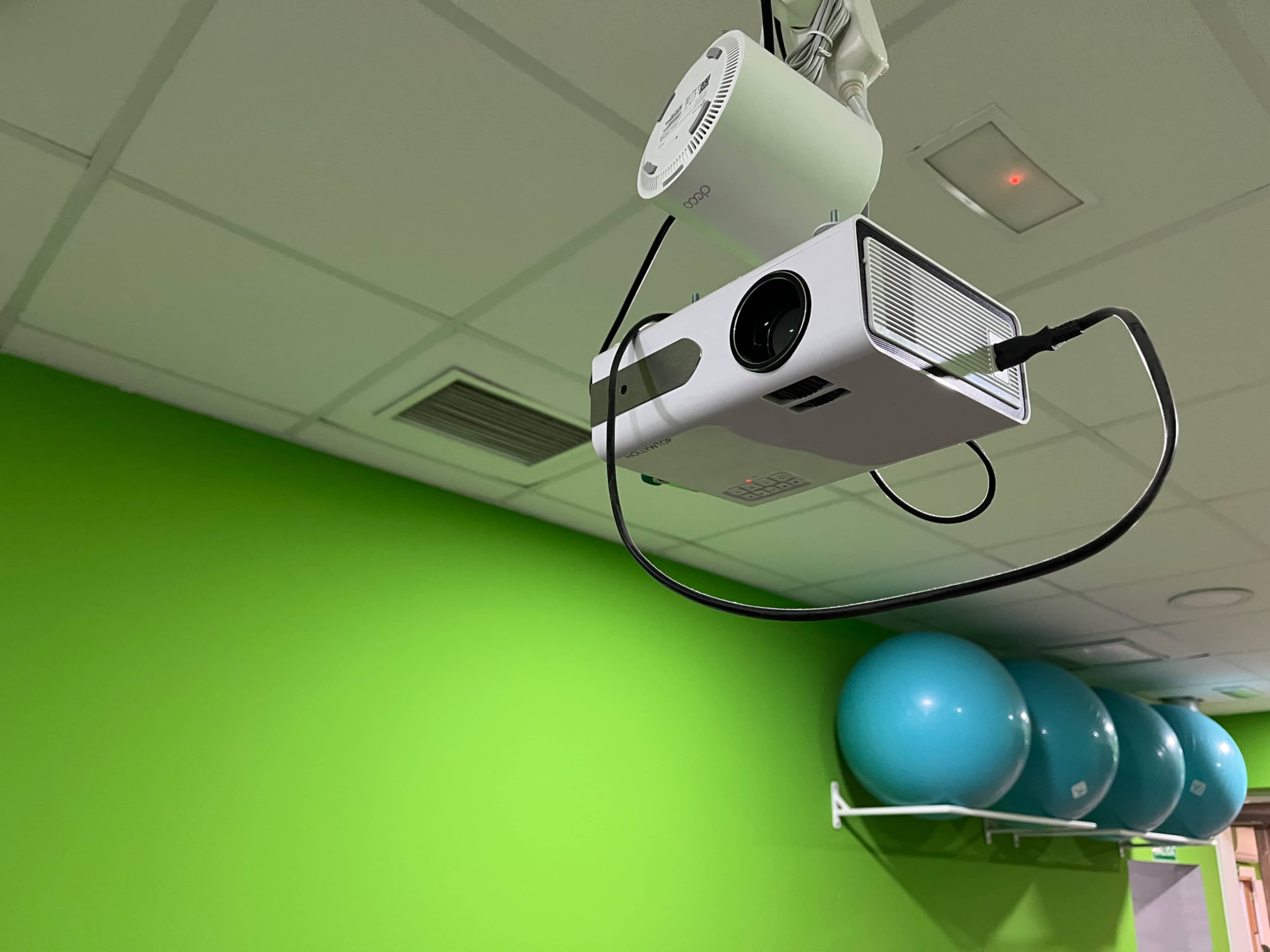 A ceiling-mounted projector is positioned in front of a bright green wall, with blue exercise balls arranged on a shelf nearby.