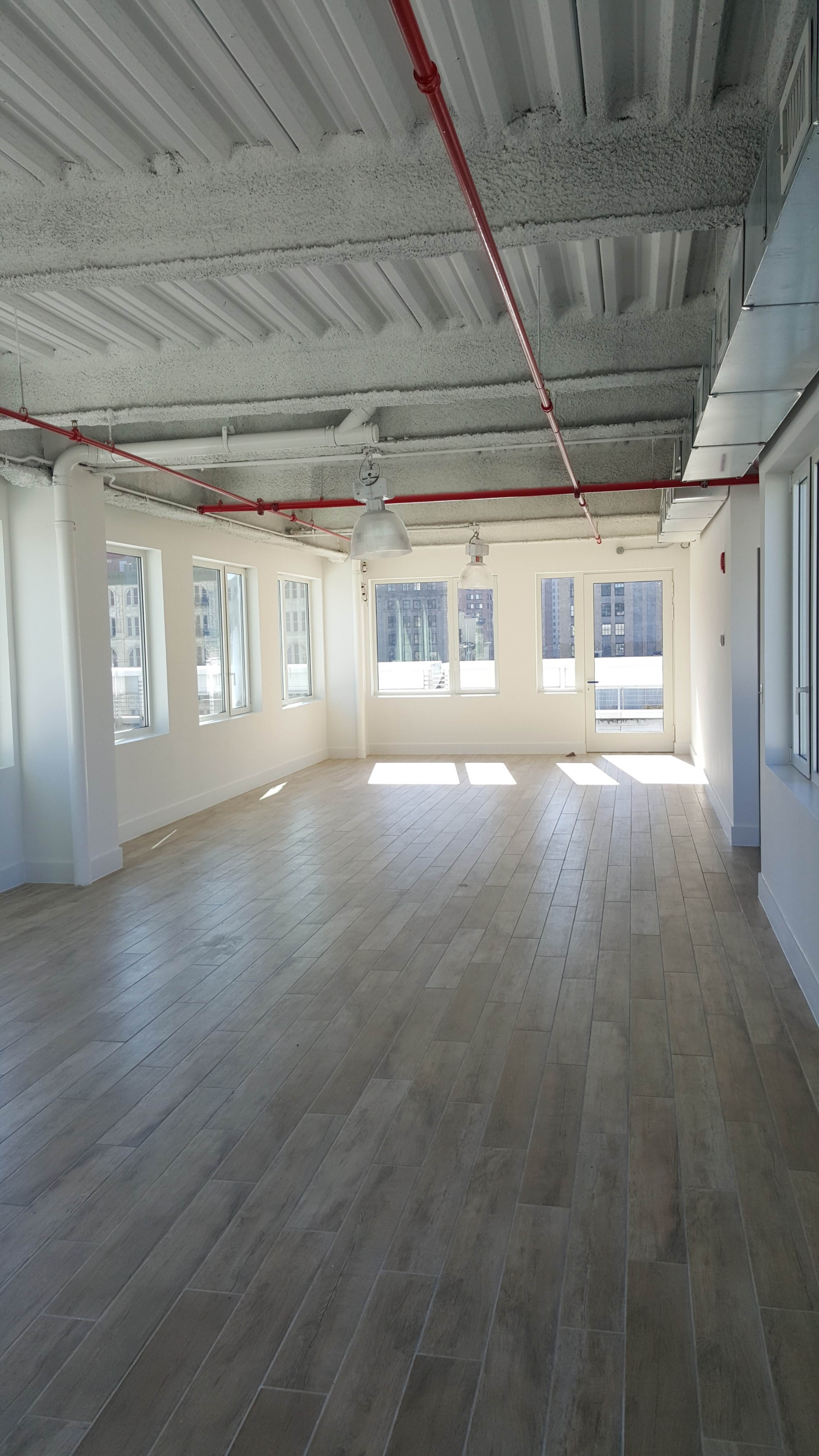 A spacious, empty office interior features large windows that allow natural light to fill the room, with exposed red piping on the ceiling.