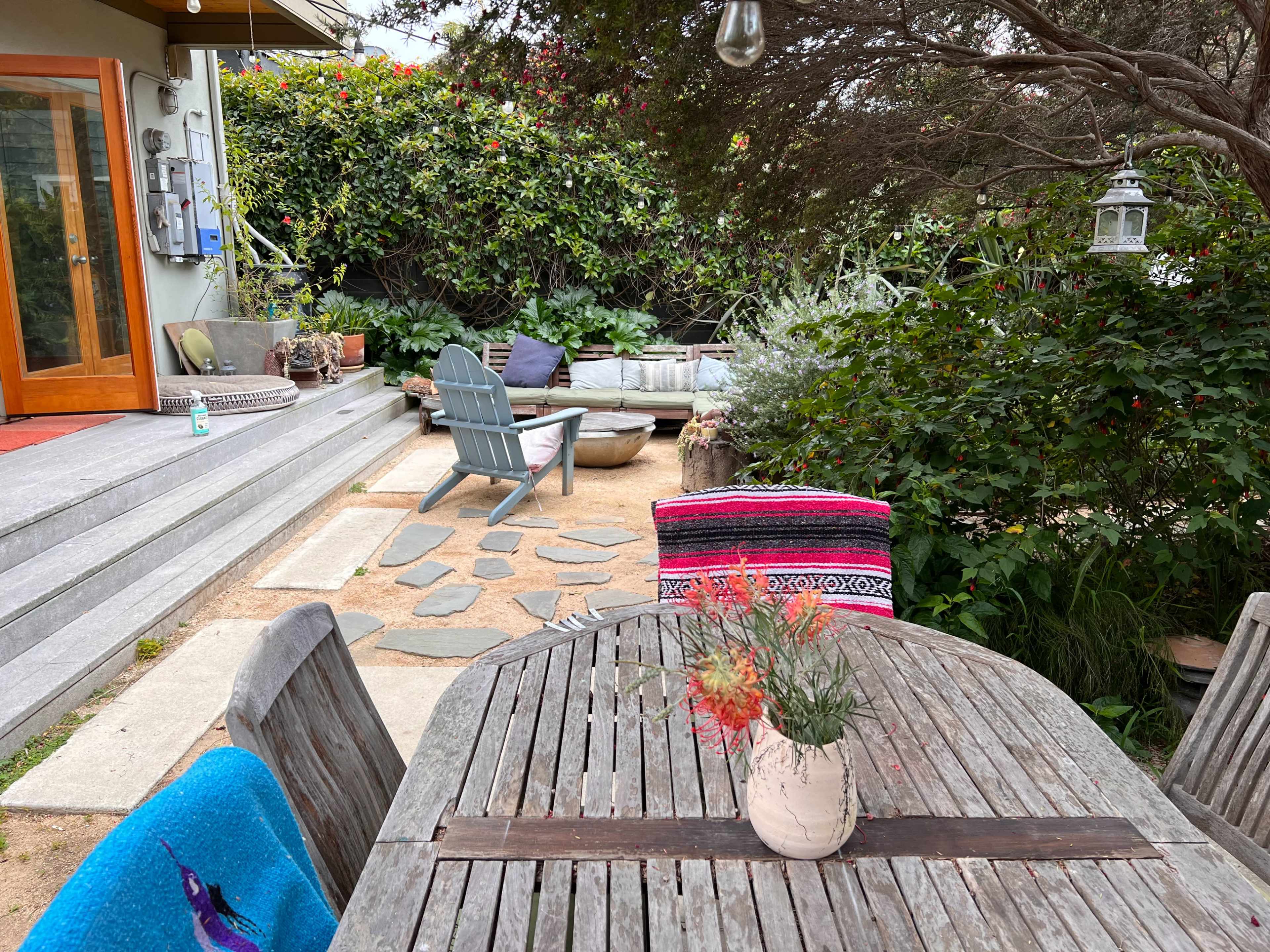 A wooden table with a vase of flowers sits in the foreground of a garden with stone pathways, surrounded by lush greenery and a cozy seating area.