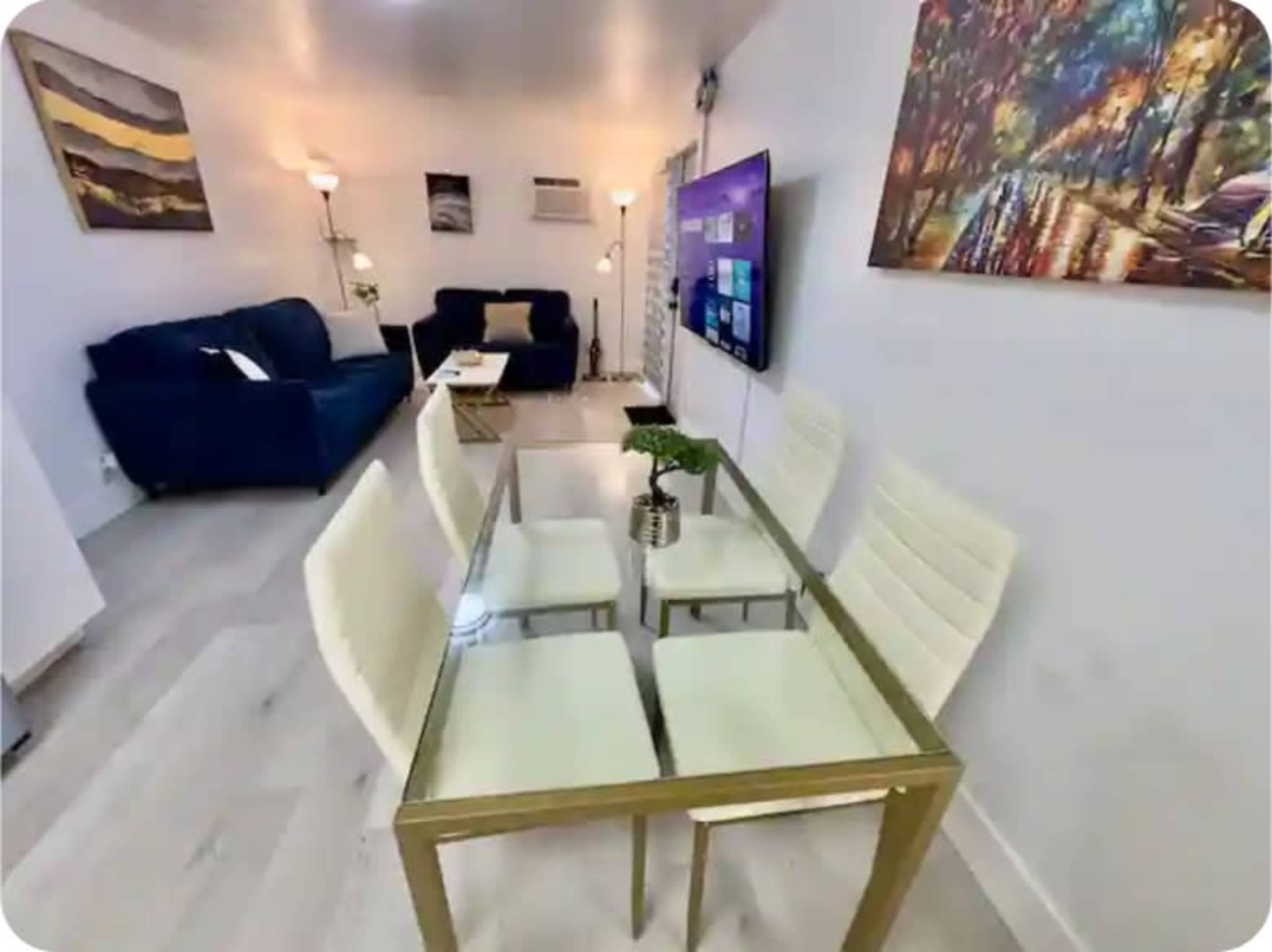 A modern living space features a glass dining table with four white chairs, a blue sofa, and a wall-mounted TV in a brightly lit room.