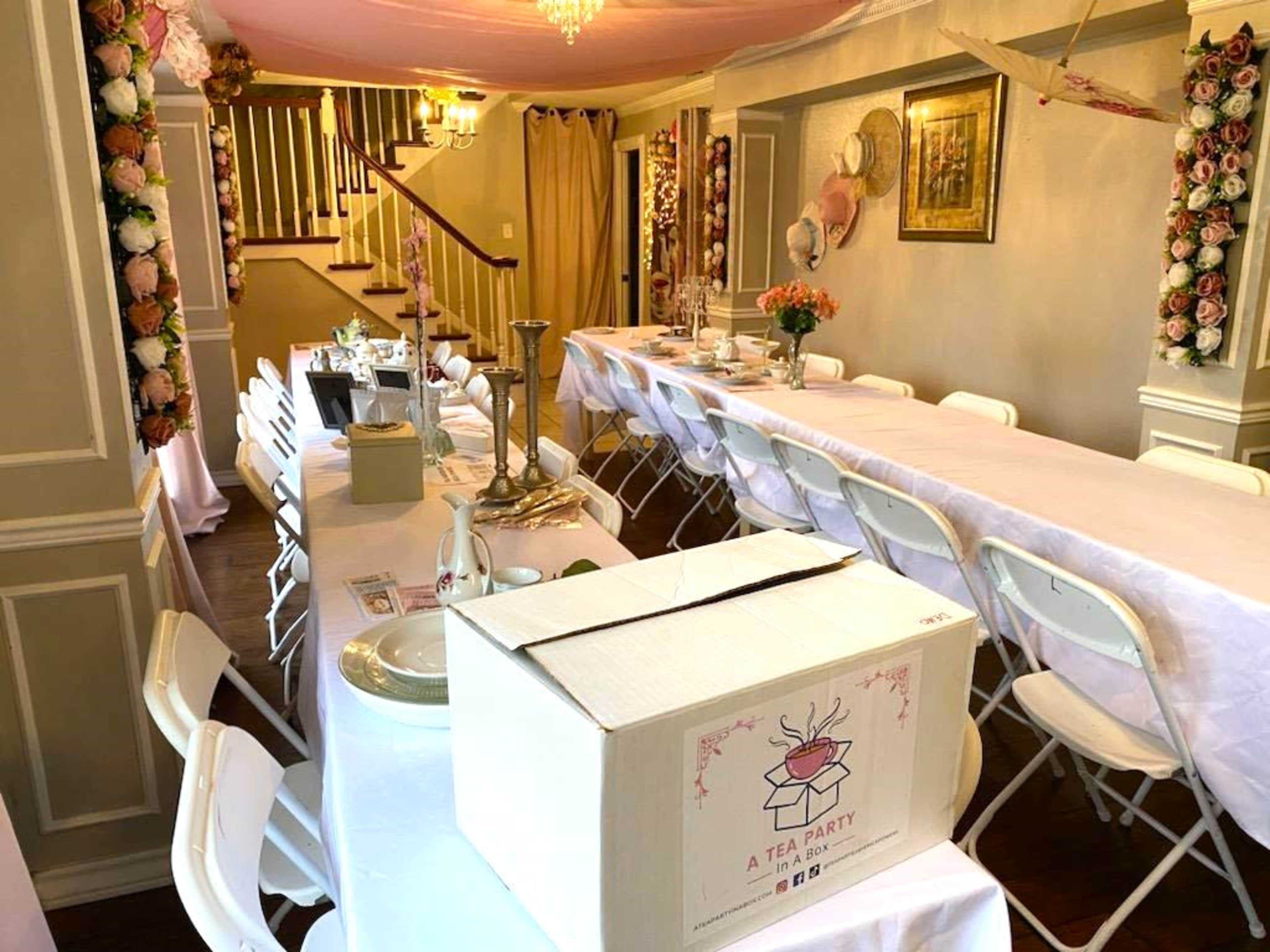 A decorated indoor space set up for a tea party, featuring long tables with white tablecloths and floral arrangements, alongside a decorative box labeled "A TEA PARTY."