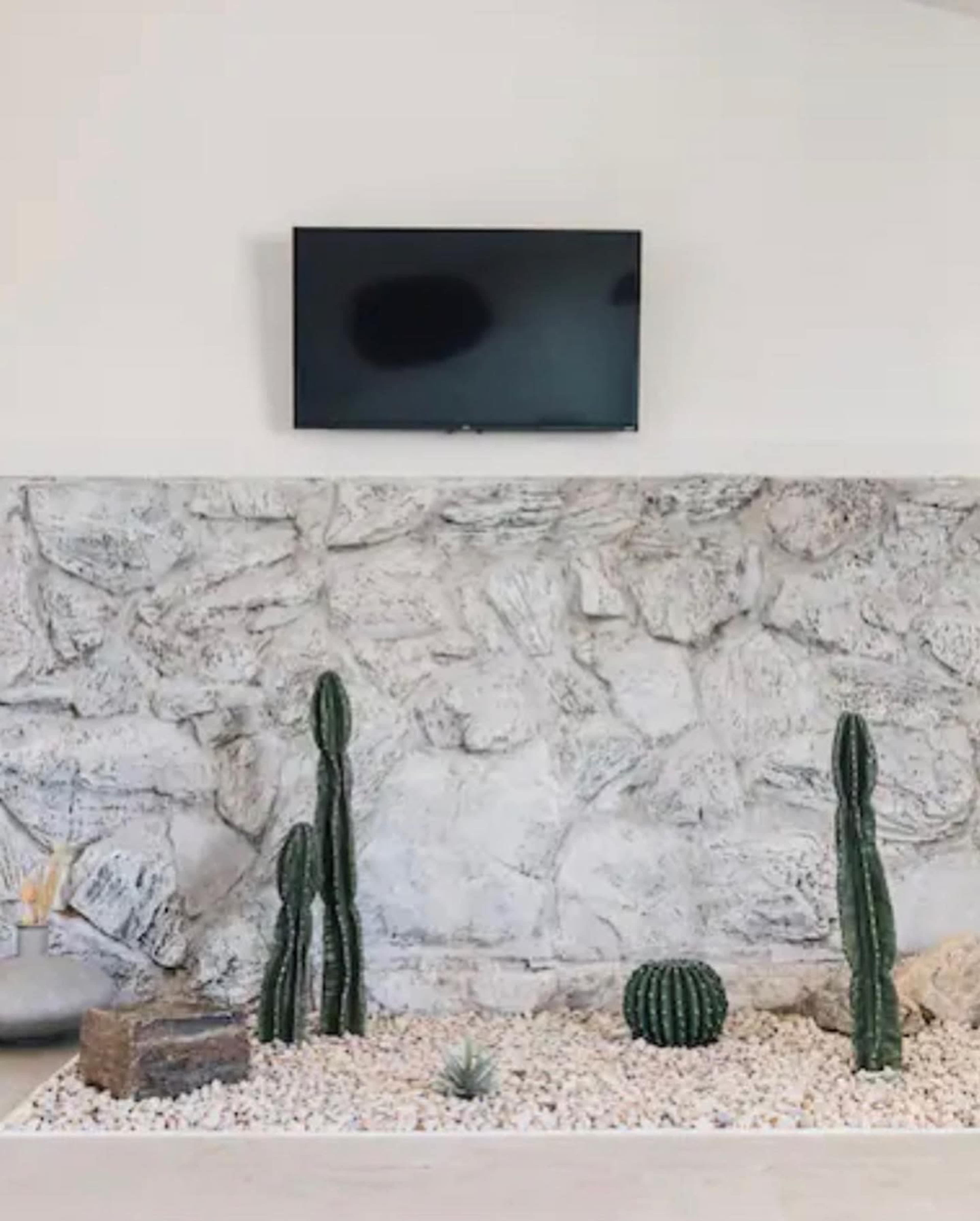 A flat-screen television is mounted on a stone wall with several cacti and white pebbles arranged at its base.
