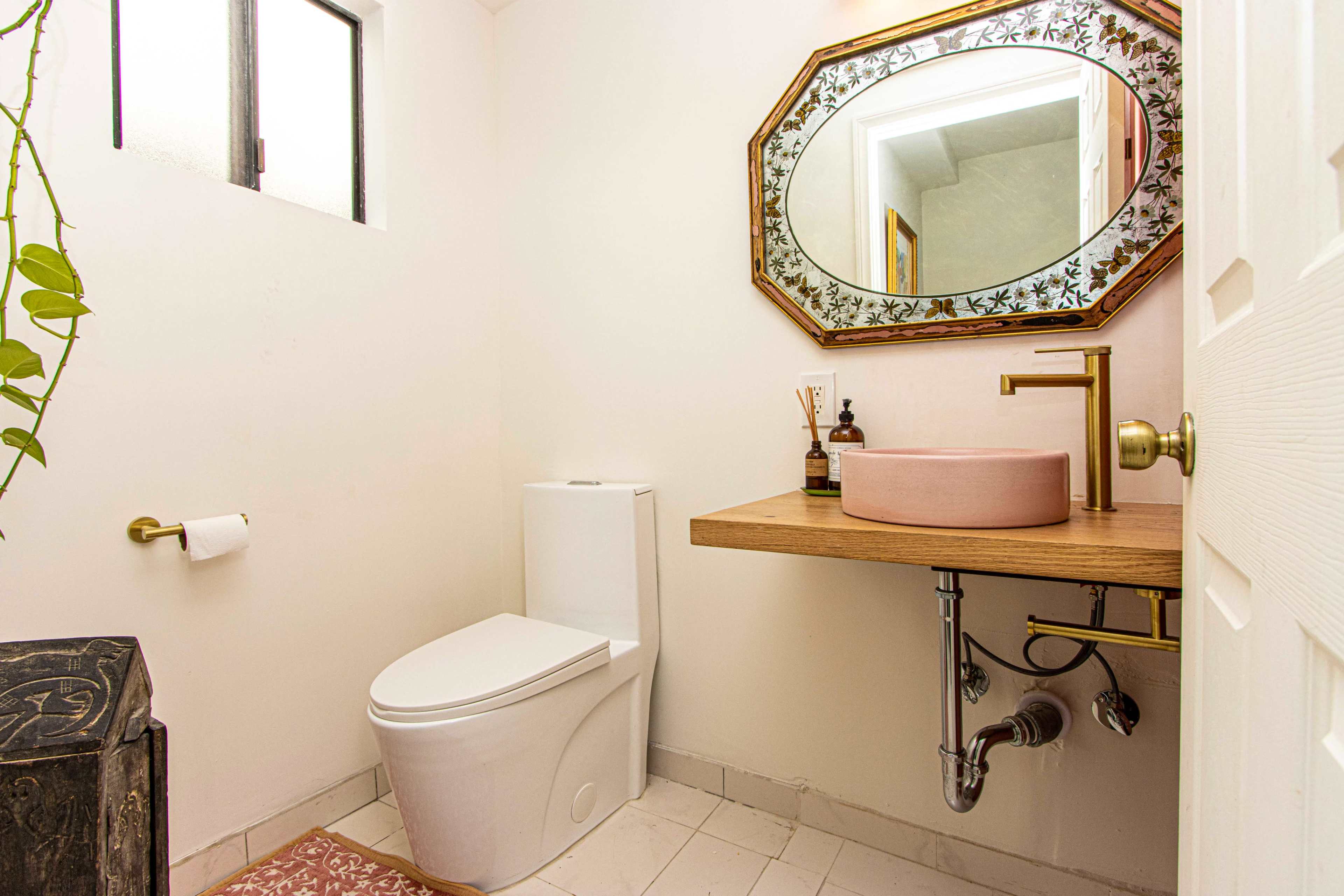 The image shows a small bathroom with a modern toilet, a pink sink on a wooden countertop, and a decorative mirror on the wall.