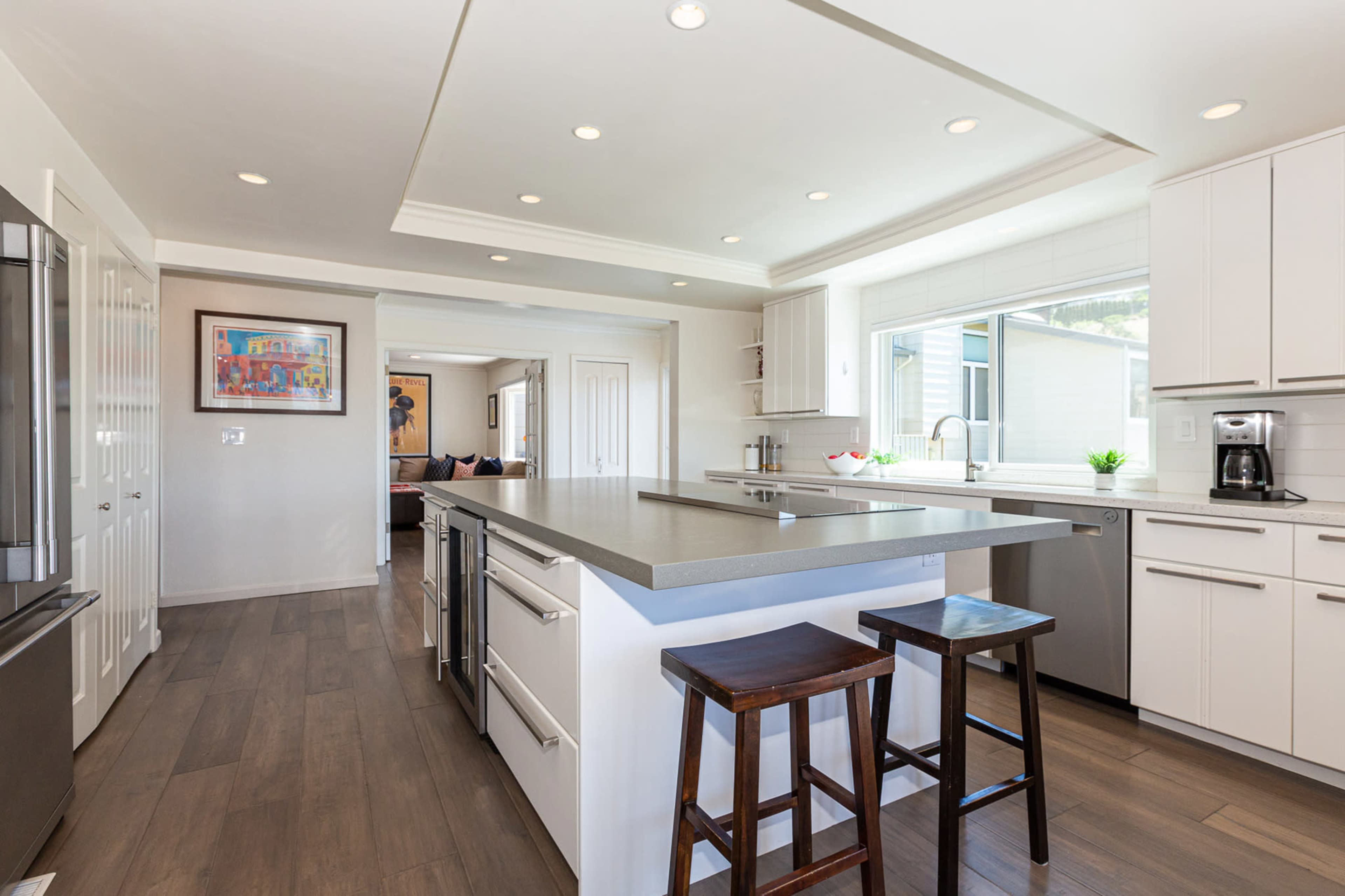A modern kitchen features white cabinetry, a central island with seating, and large windows providing natural light.