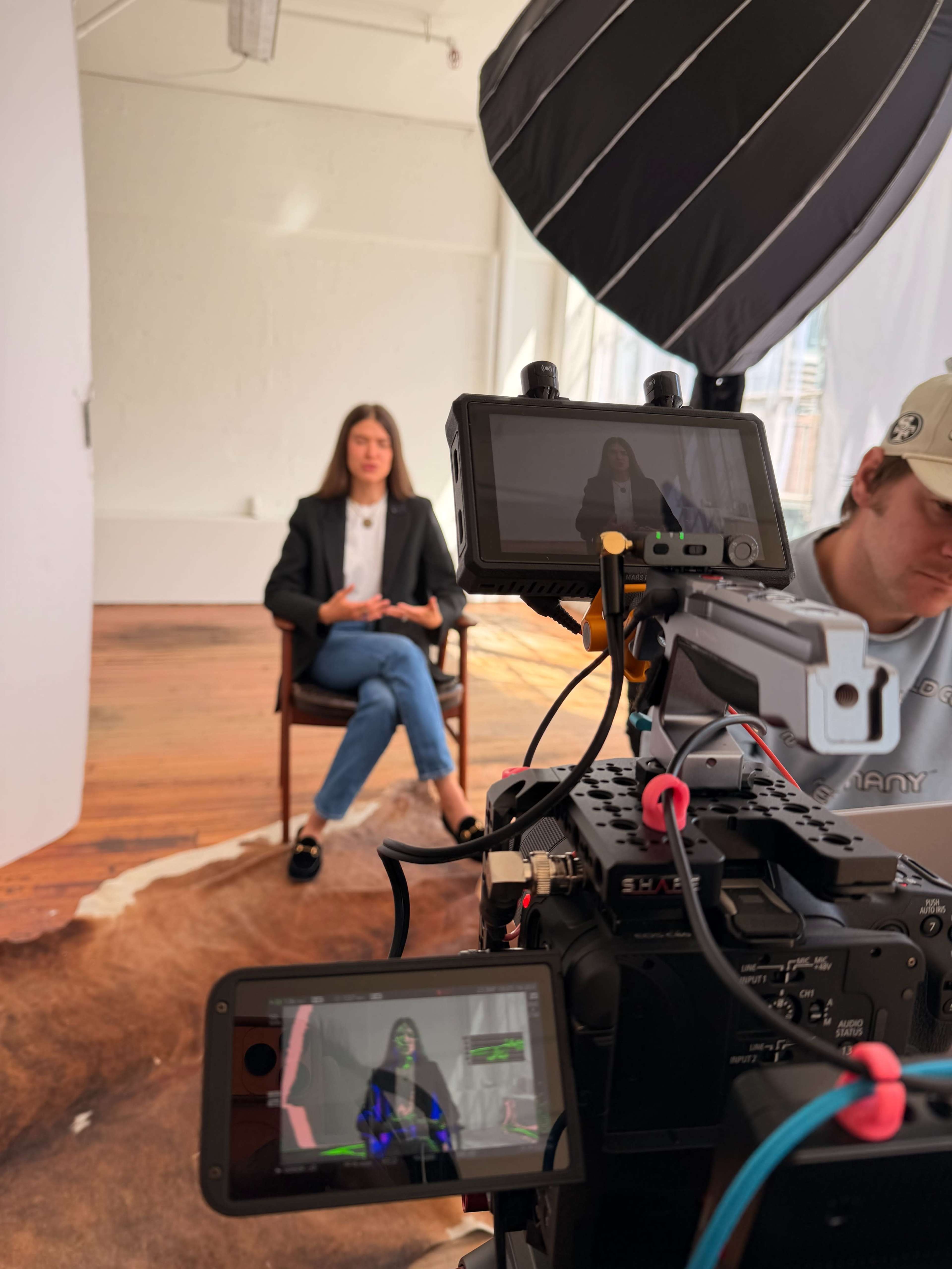 A woman sits on a chair in front of a camera setup with a bright light and backdrop during a photoshoot.