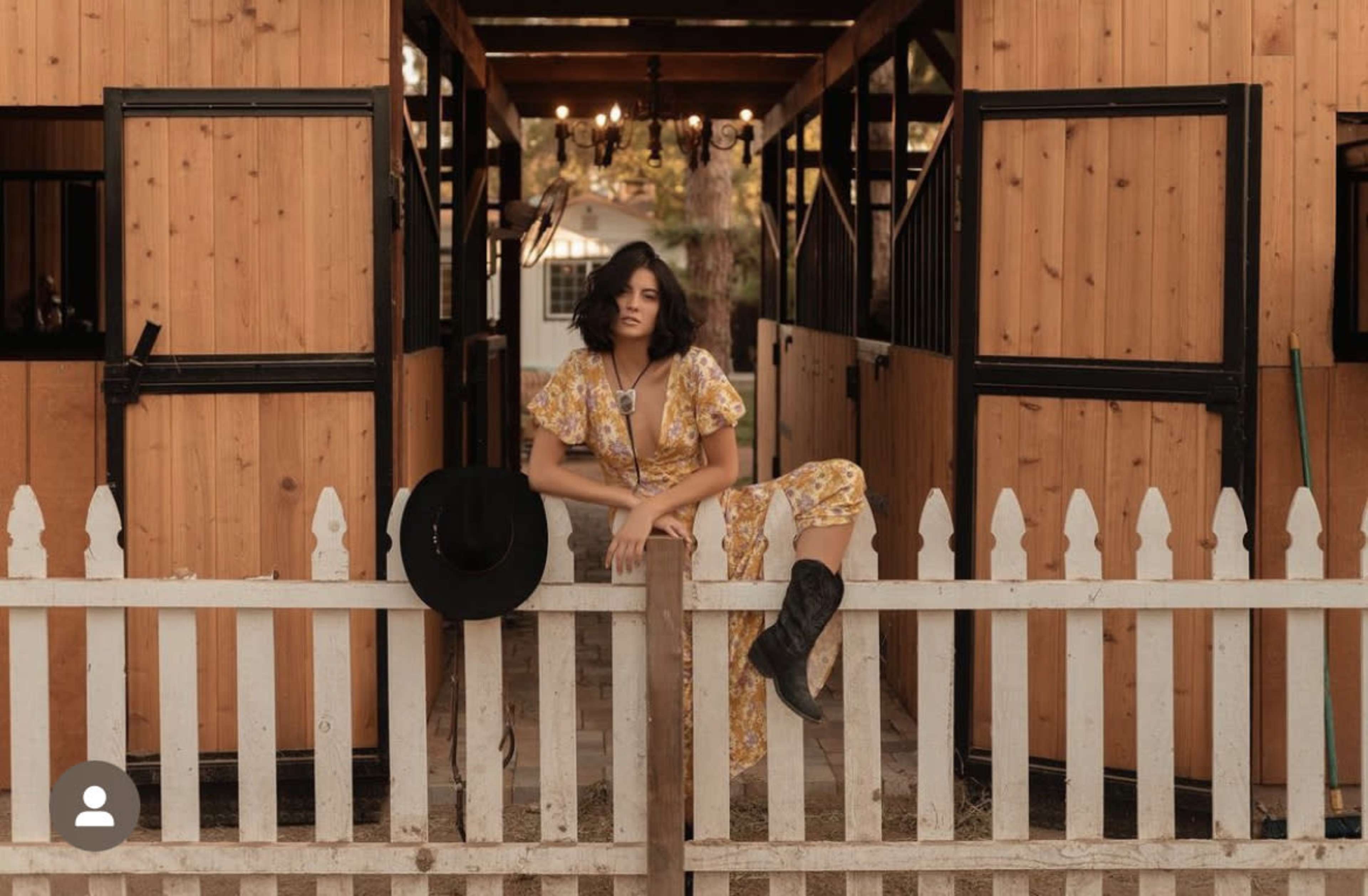 A woman in a floral outfit sits on a white picket fence between two horse stables.