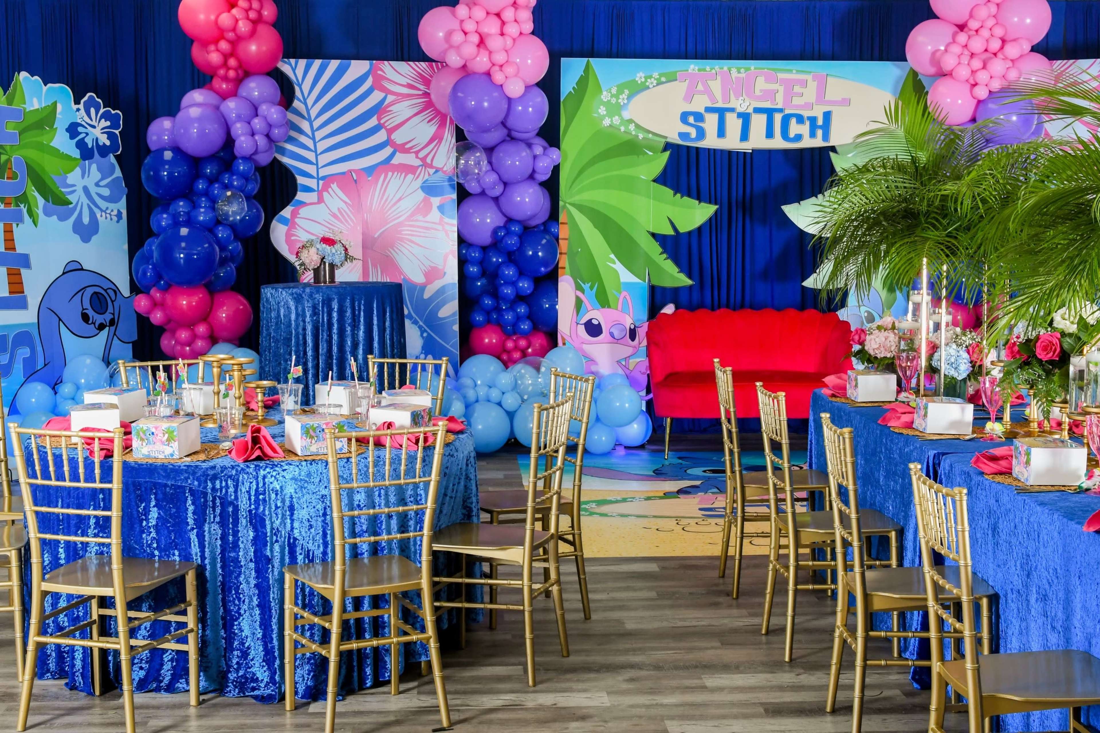 A party setup featuring two round tables with blue tablecloths, gold chairs, colorful balloon decorations, and a backdrop with the words "Angel Stitch" along with tropical themed elements.