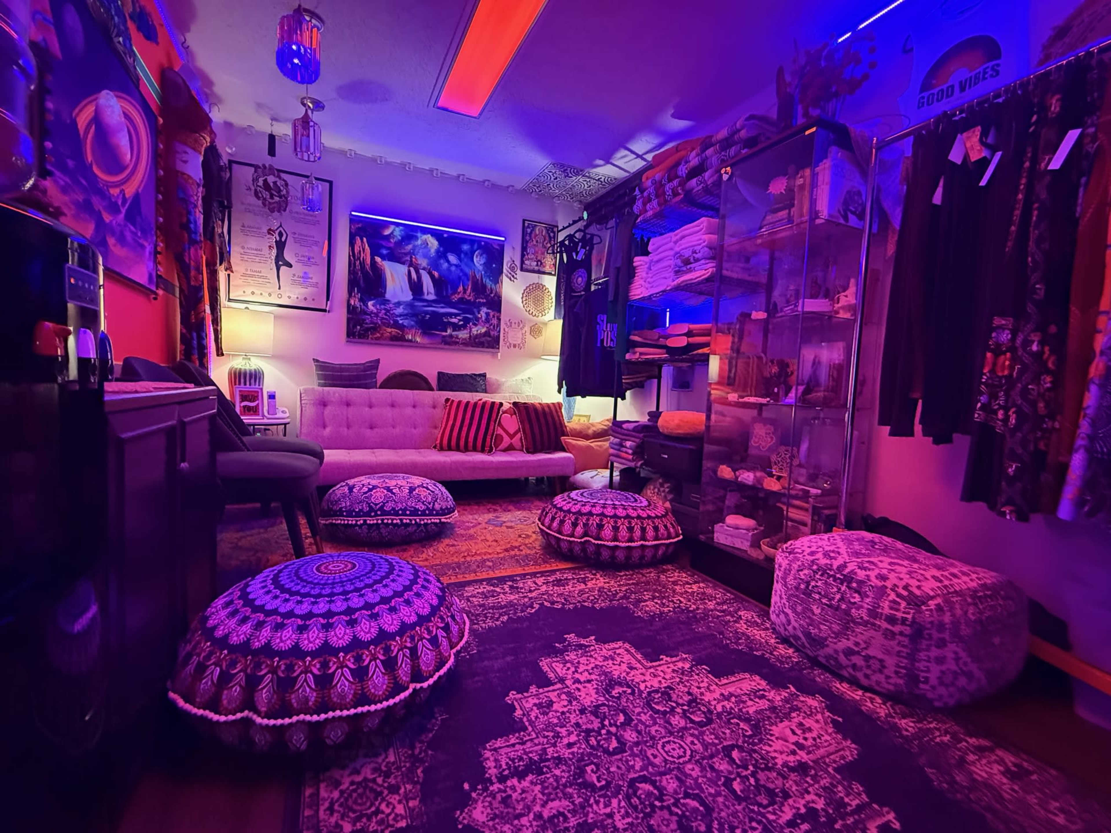 The room features a cozy seating area with a sofa, colorful cushions, poufs, and shelves displaying various items, all illuminated by purple and blue lighting.