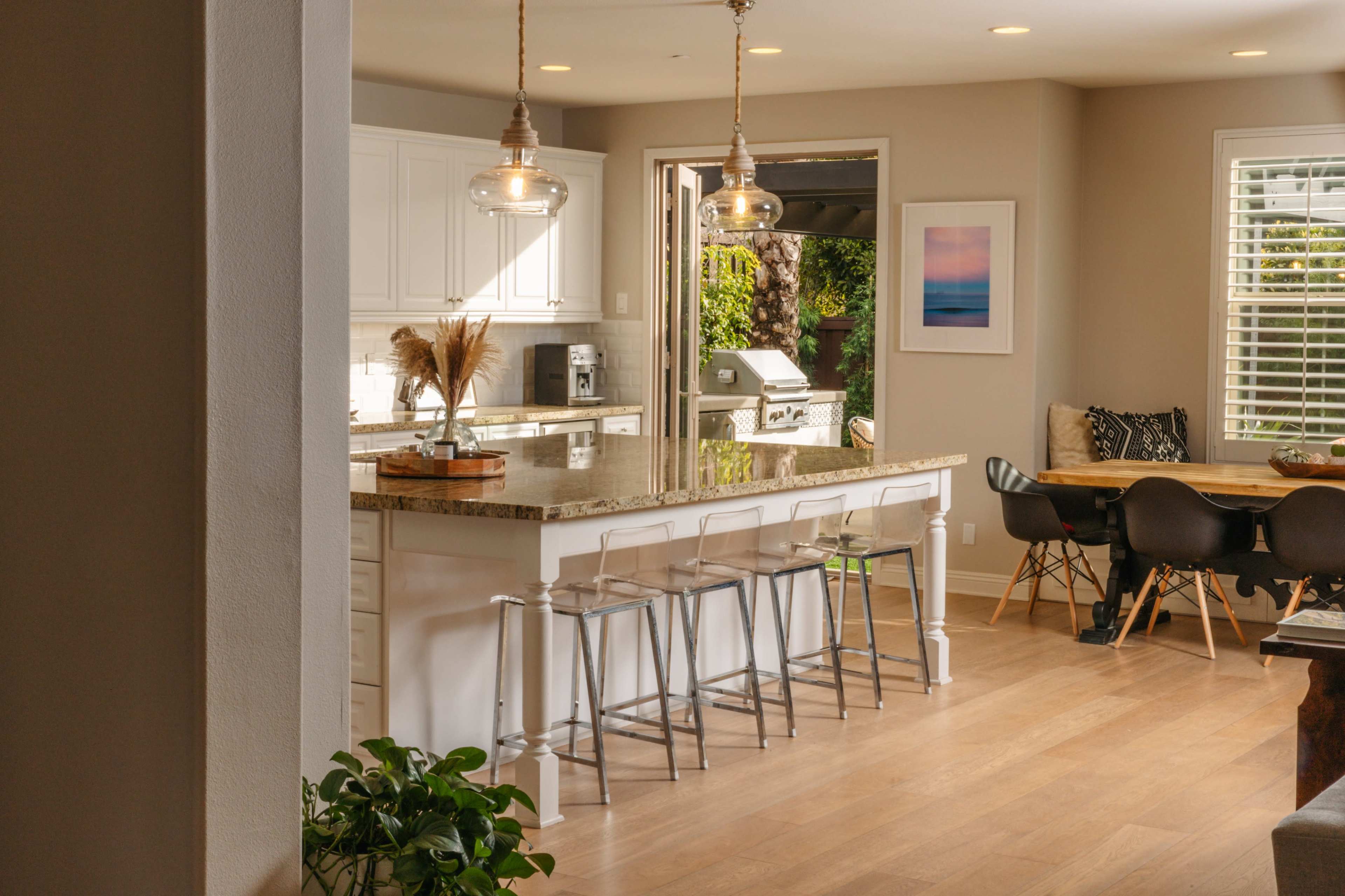 A modern kitchen features a large island with bar stools, a dining area, and natural light coming through the open door, revealing an outdoor space.
