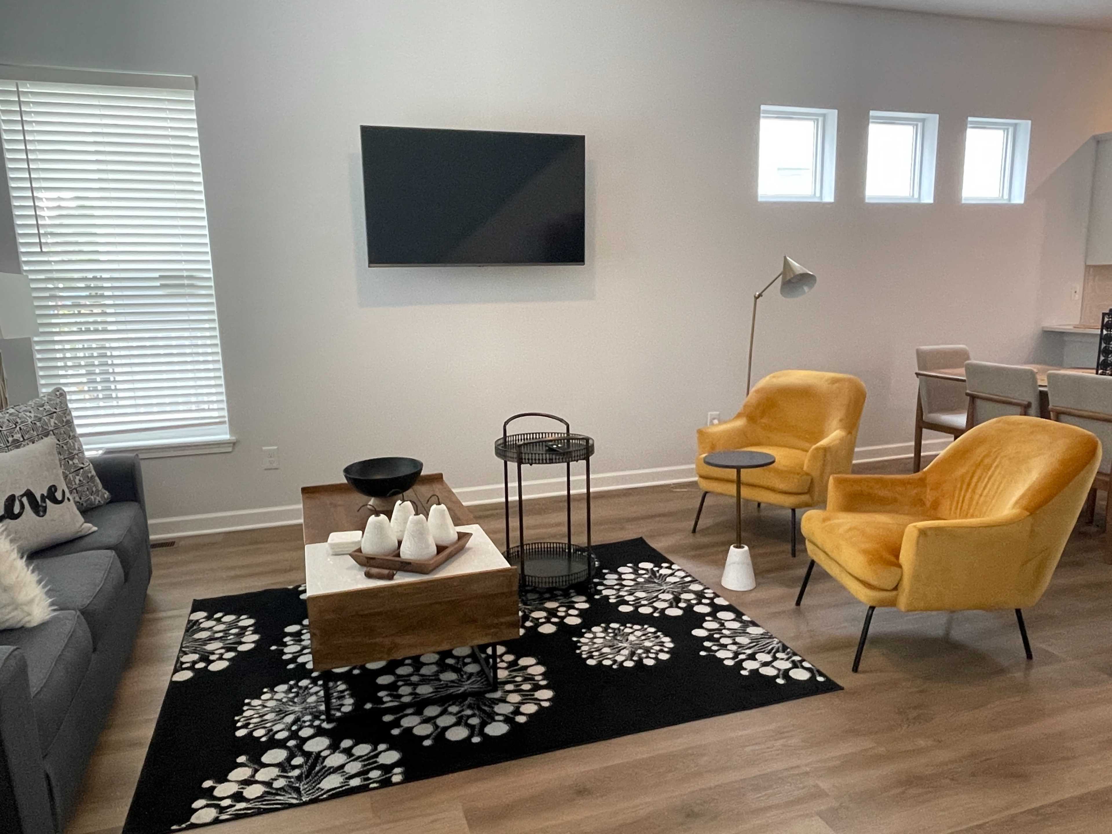 The living room features a gray sofa, two yellow armchairs, a wooden coffee table with decorative items, and a television mounted on the wall.