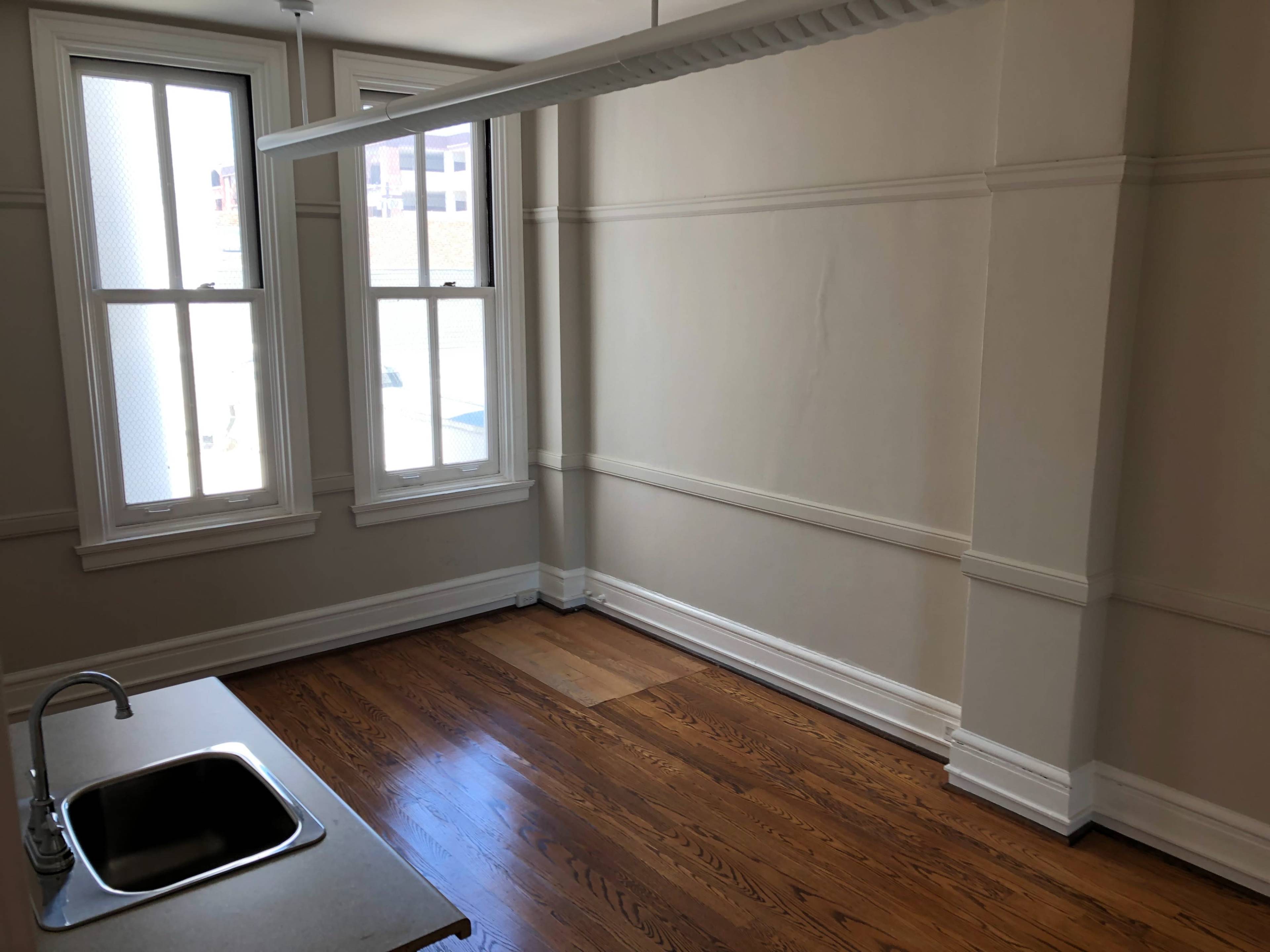 An empty room with wooden flooring, two windows, and a small kitchenette in one corner.