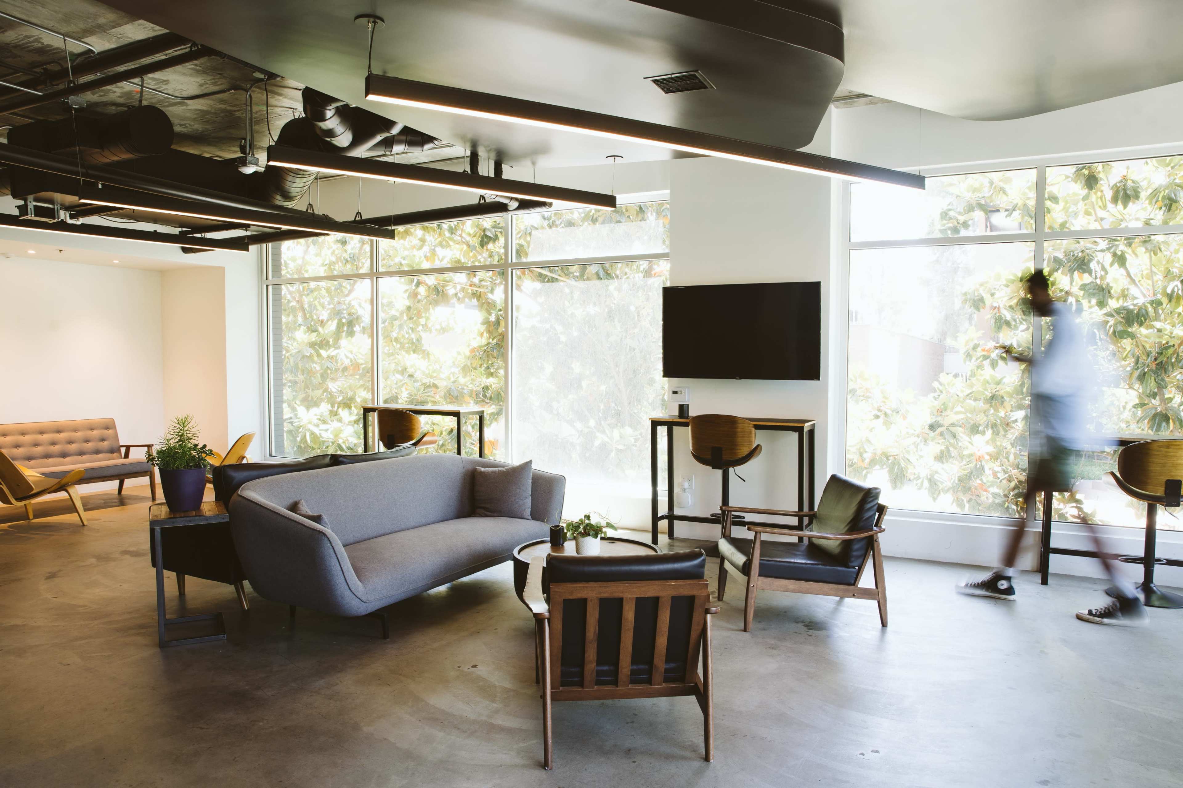 A modern lounge area with large windows, a gray sofa, wooden chairs, and a television mounted on the wall.