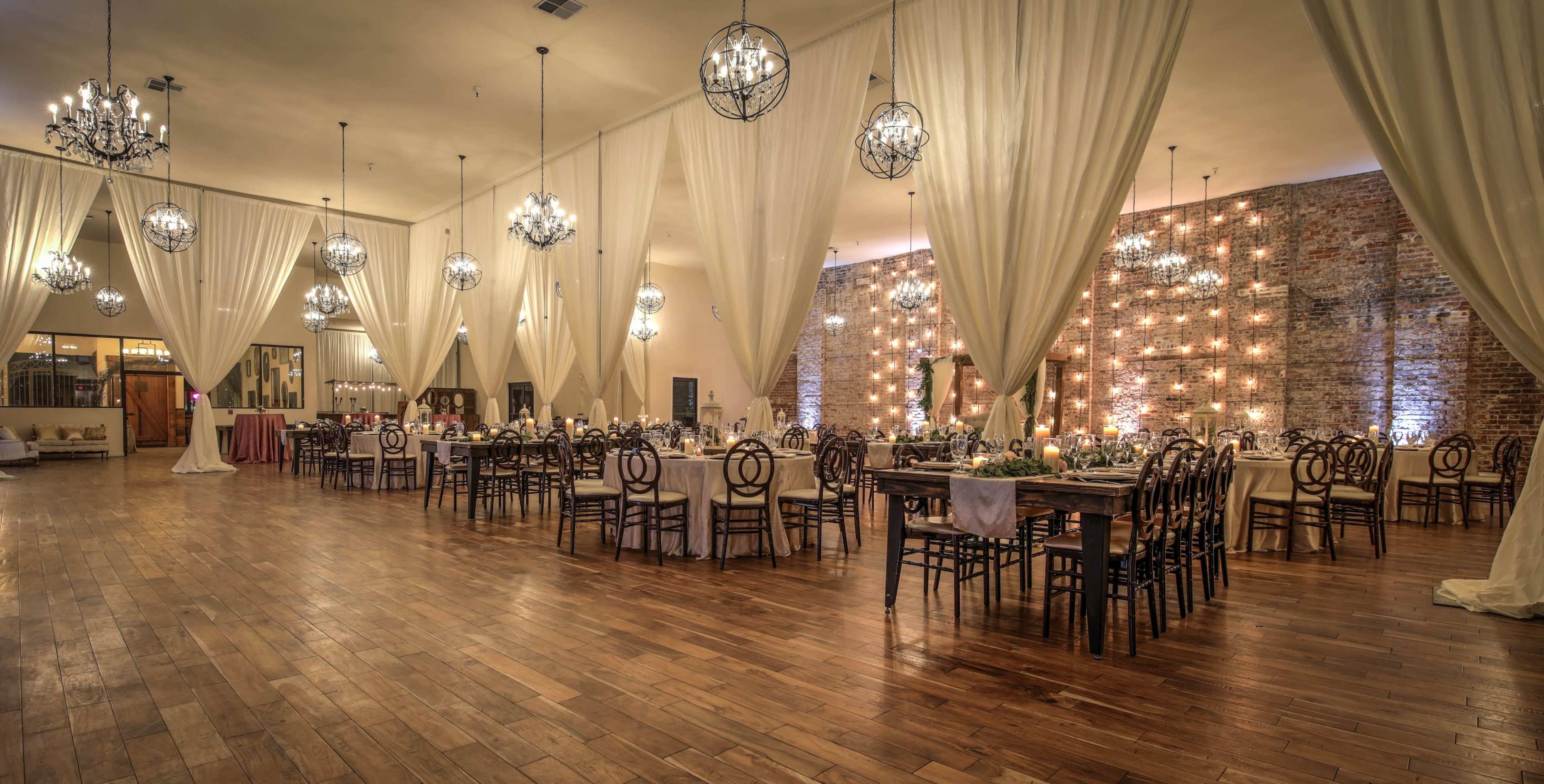 A spacious event hall featuring wooden floors, long dining tables, and chandeliers, with draped fabric along the ceiling and a brick wall adorned with lights.