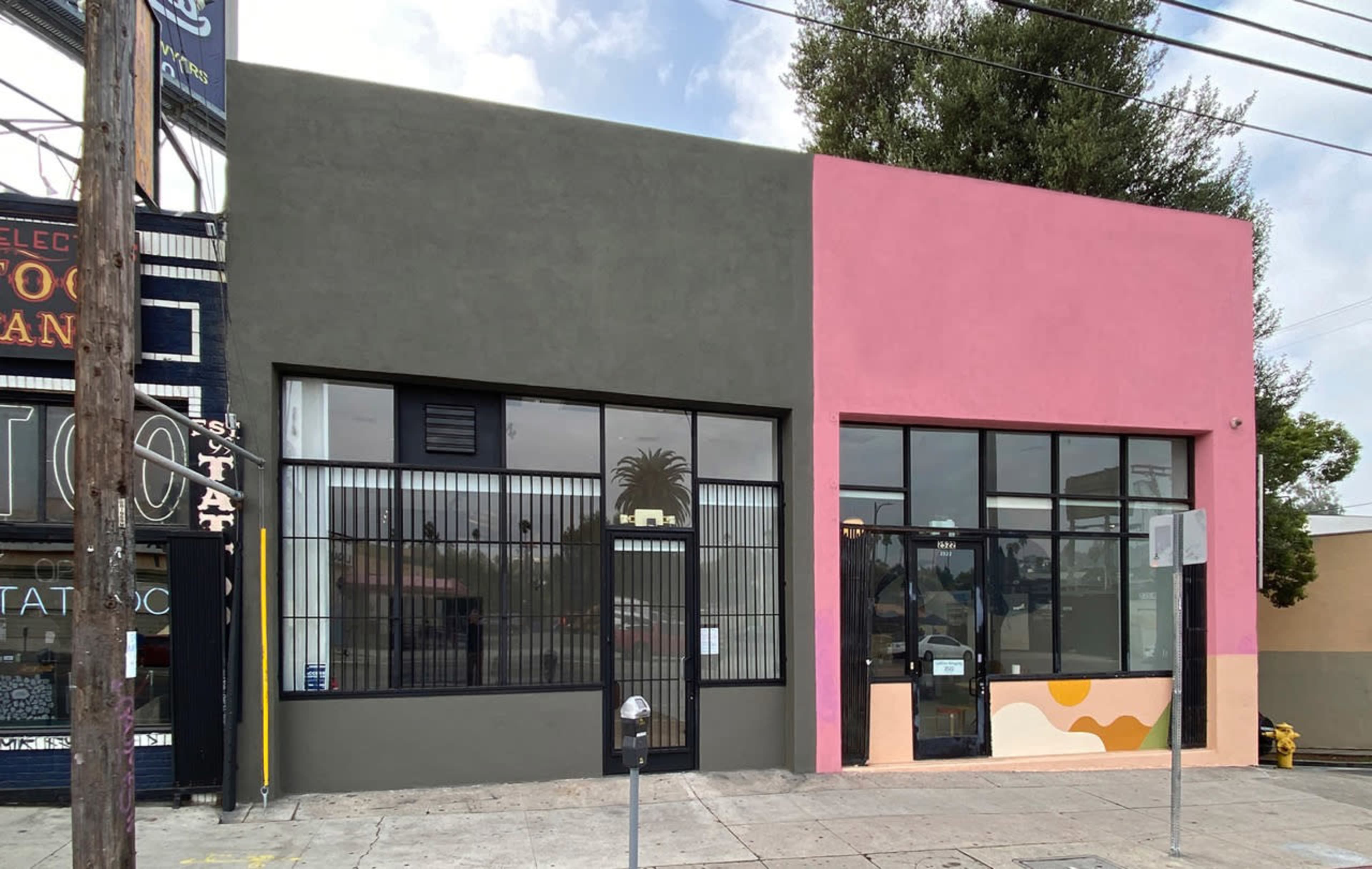 The image shows a two-toned building with a green left side and a pink right side, featuring large windows and a sidewalk in front.