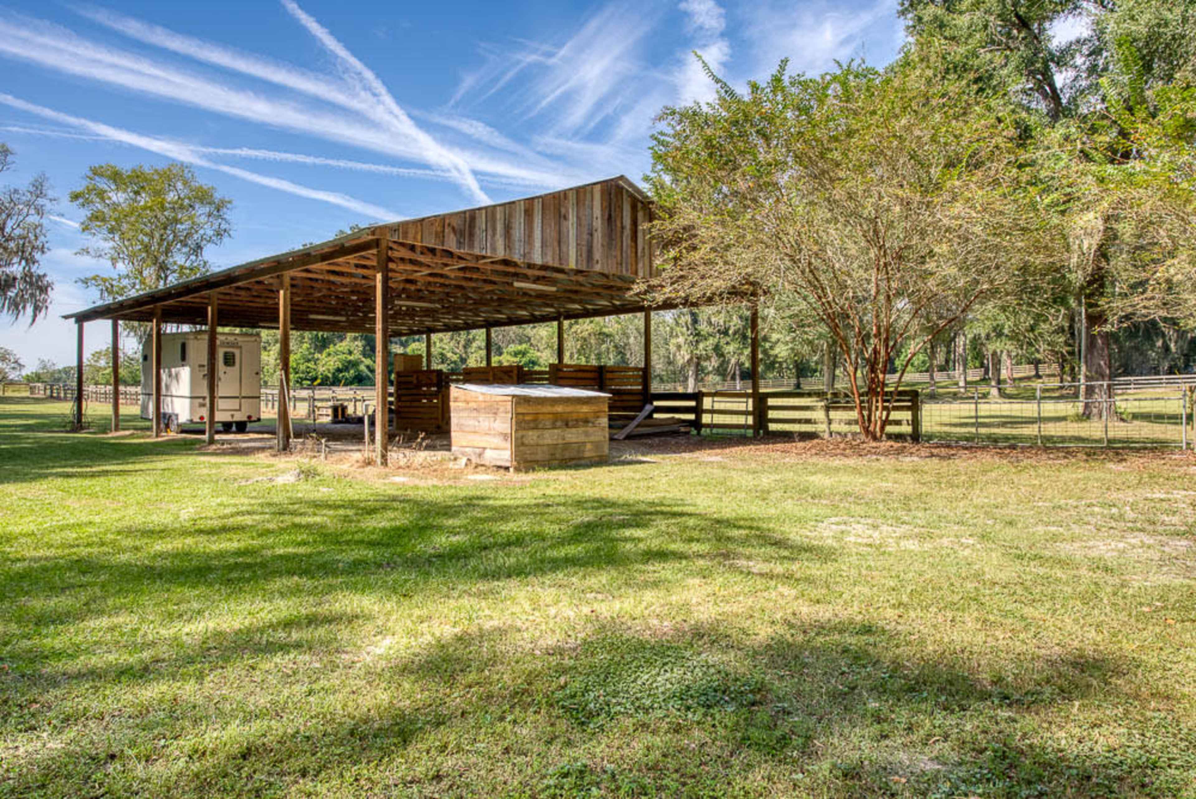 The image shows a spacious open shelter with wooden supports, located in a grassy area surrounded by trees and a fence.