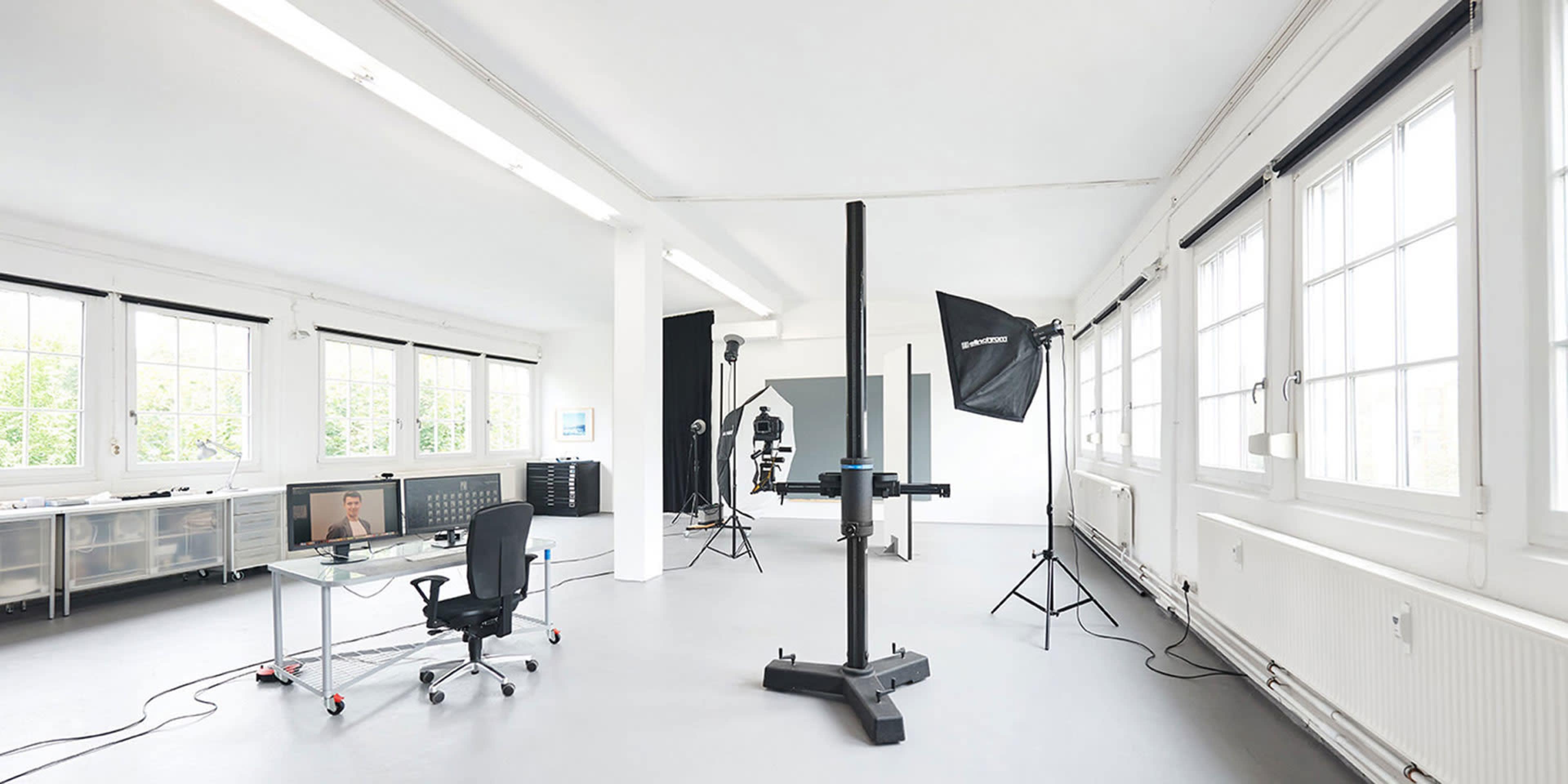 The image shows a spacious photography studio featuring a large backdrop, various lighting equipment, a desk with a computer and chair, and several windows.