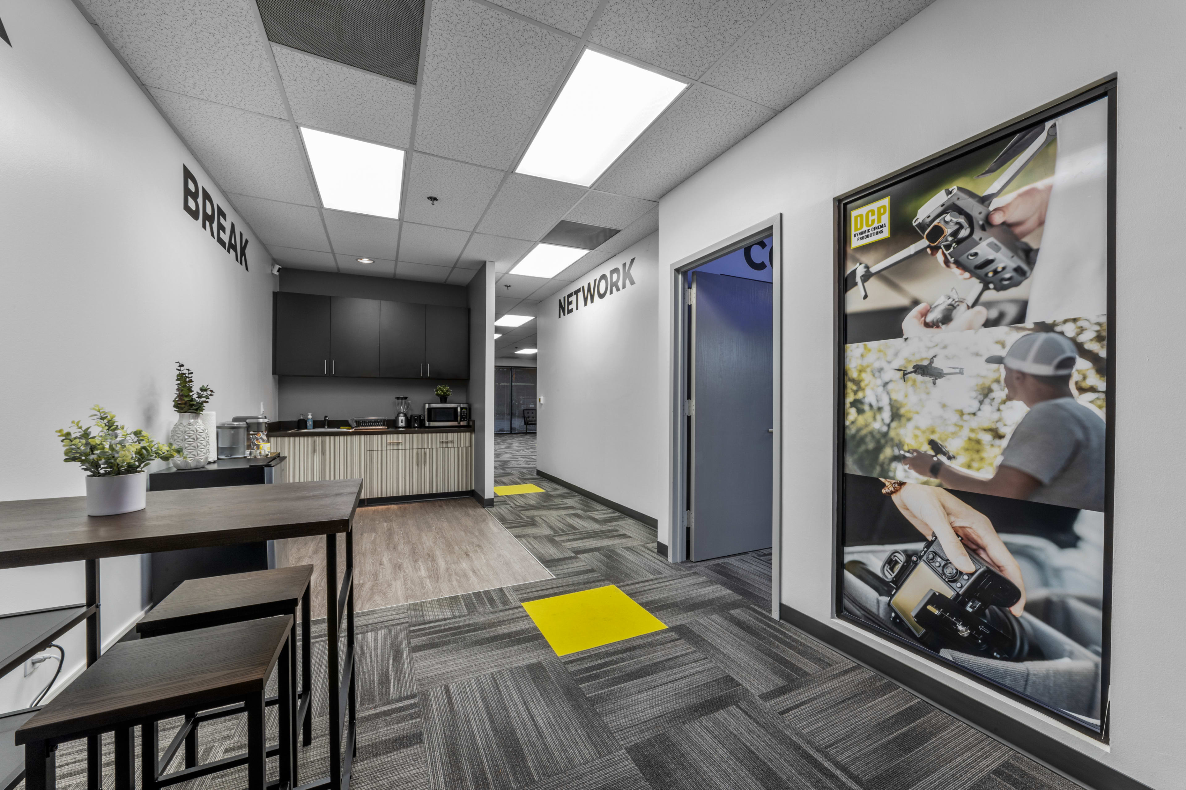 A modern office hallway features a kitchen area on one side and a large poster of a photographer on the other, with yellow and gray floor tiles.